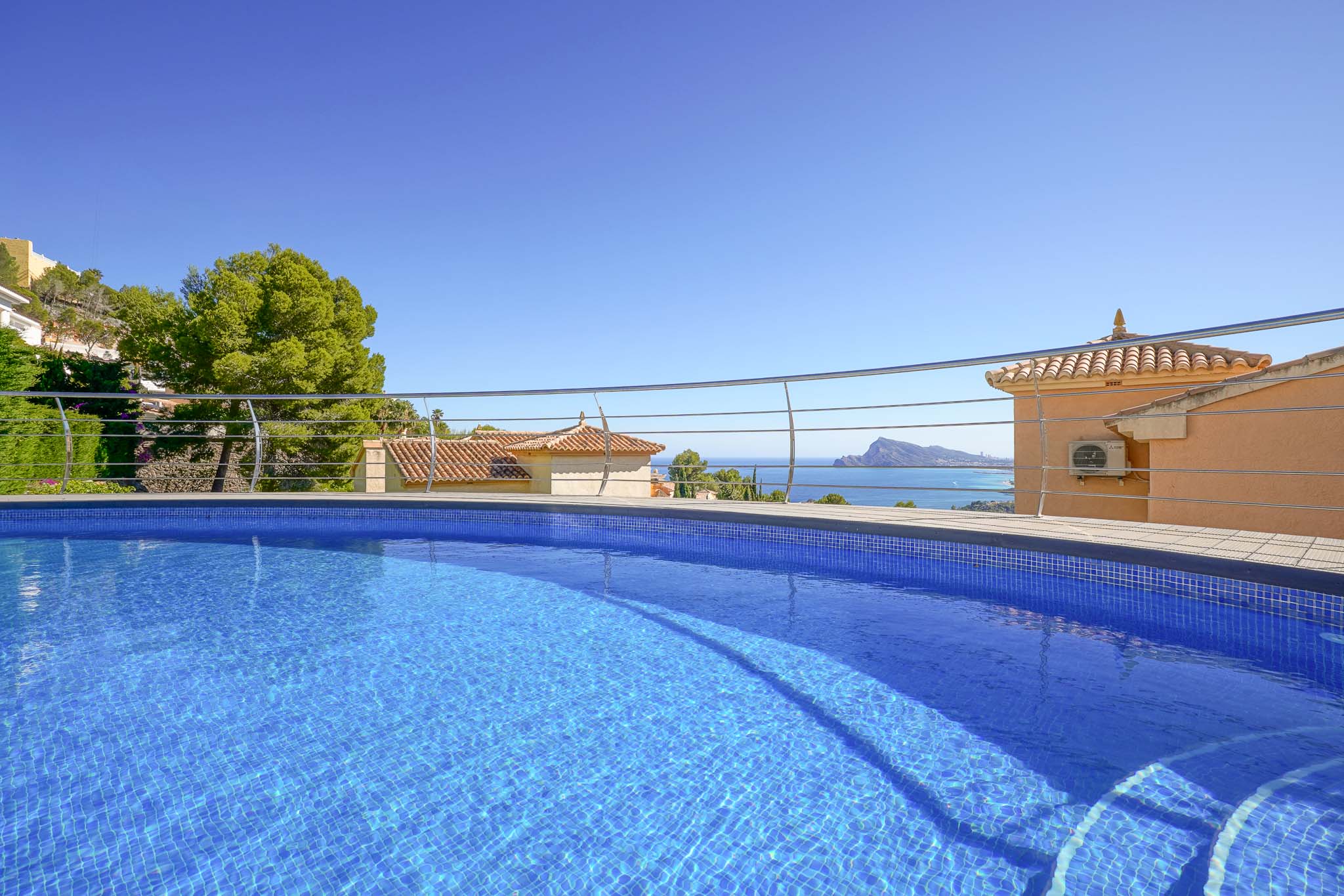 Blauer Mosaik-Swimmingpool mit integrierten Stufen und modernem Geländer, der einen Panoramablick auf das Meer, traditionelle Dächer und die Küste bietet.