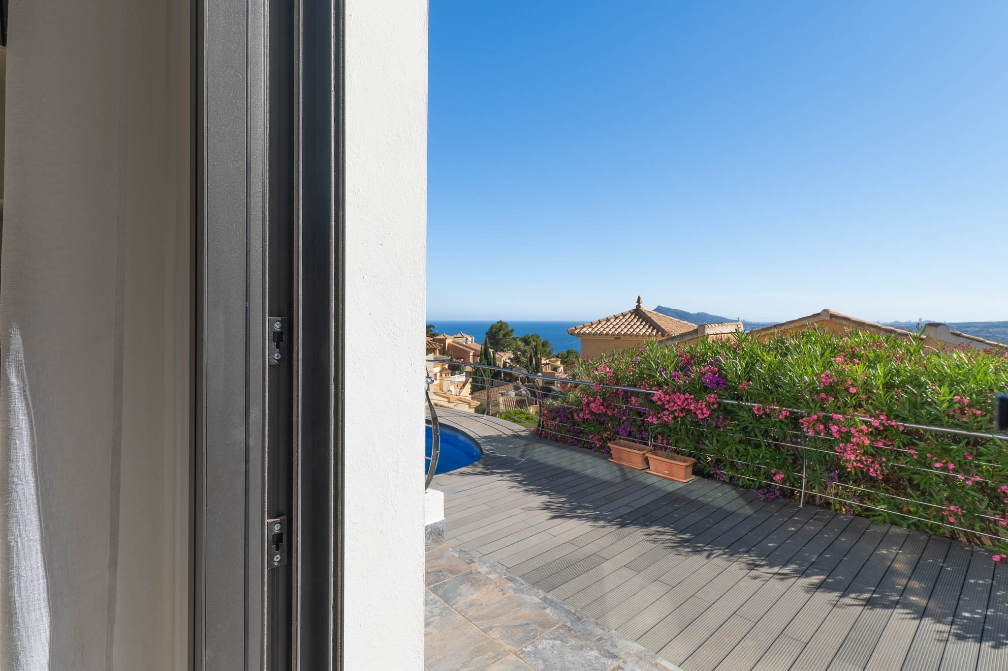Blick von der Schiebetür auf die Terrasse mit WPC-Dielen, Poolausschnitt und Edelstahlgeländer. Im Hintergrund sind das Mittelmeer und traditionelle Dächer zu sehen.