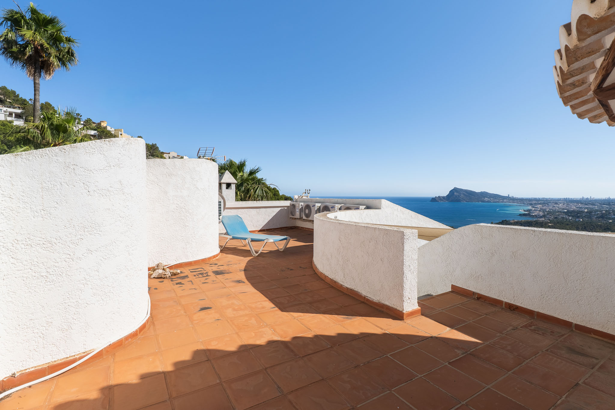 Dachterrasse mit Terrakottafliesen und weißen Strukturwänden, die einen Panoramablick auf das Meer und die Küste bietet. Eine blaue Sonnenliege lädt zum Entspannen ein.
