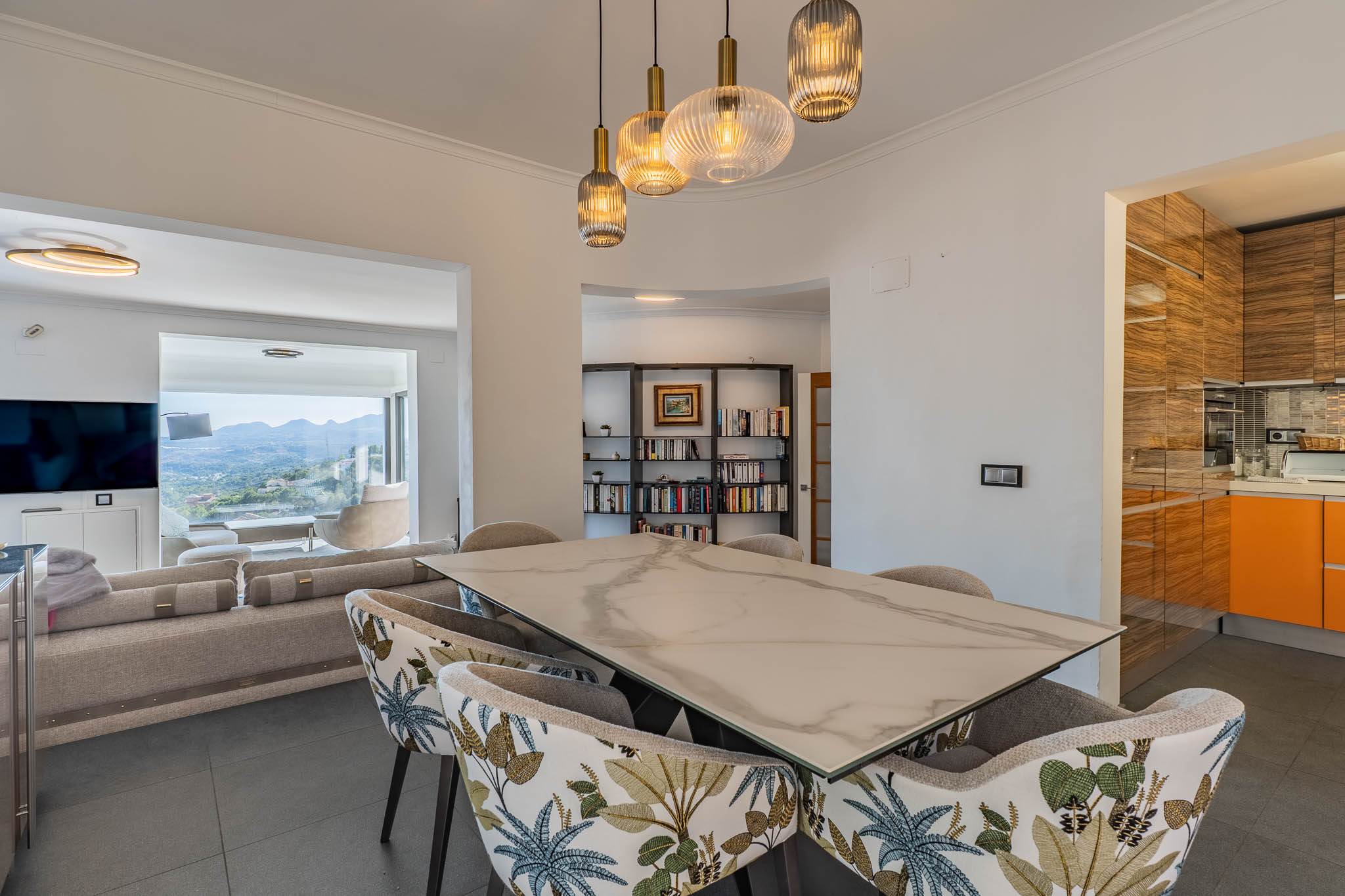 Design-Essbereich mit Marmor-Effekt-Tisch und gemusterten Stühlen, beleuchtet von modernen Pendelleuchten. Verbindet sich mit dem Wohnzimmer und bietet Panoramablicke auf die Berge.