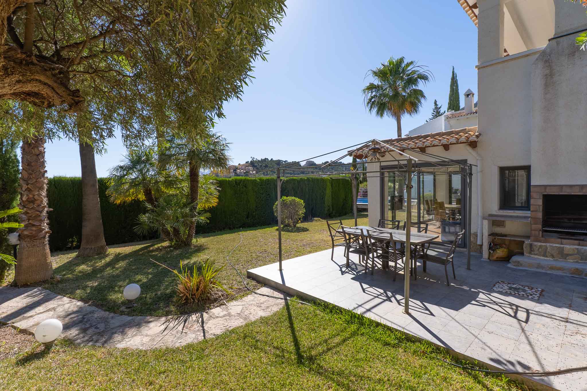 Dieser einladende Essbereich im Freien verfügt über eine geflieste Terrasse mit Metallpergola, ideal für Mahlzeiten. Ergänzt wird er durch einen gemauerten Grill und einen üppigen Garten mit Palmen und Hecke.