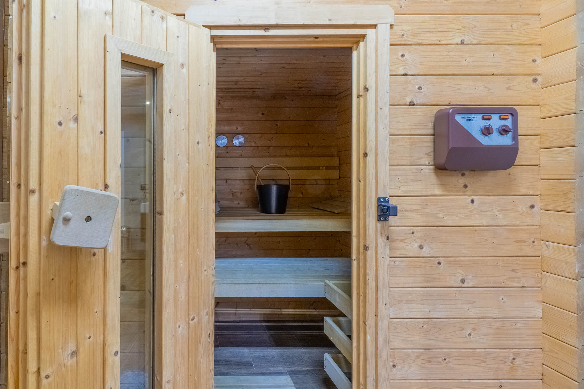 Eingang zur Sauna mit heller Holzverkleidung, Blick auf die Innenbänke, einen Eimer mit Kelle und das externe Bedienfeld.