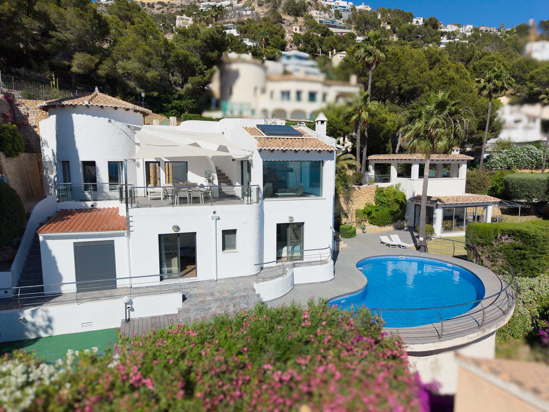 Erhöhte Ansicht der Hauptvilla mit großer oberen Terrasse, Essbereich und Markise, einem nierenförmigen Pool mit Deck und einem separaten Gästehaus, umgeben von üppiger mediterraner Landschaft.