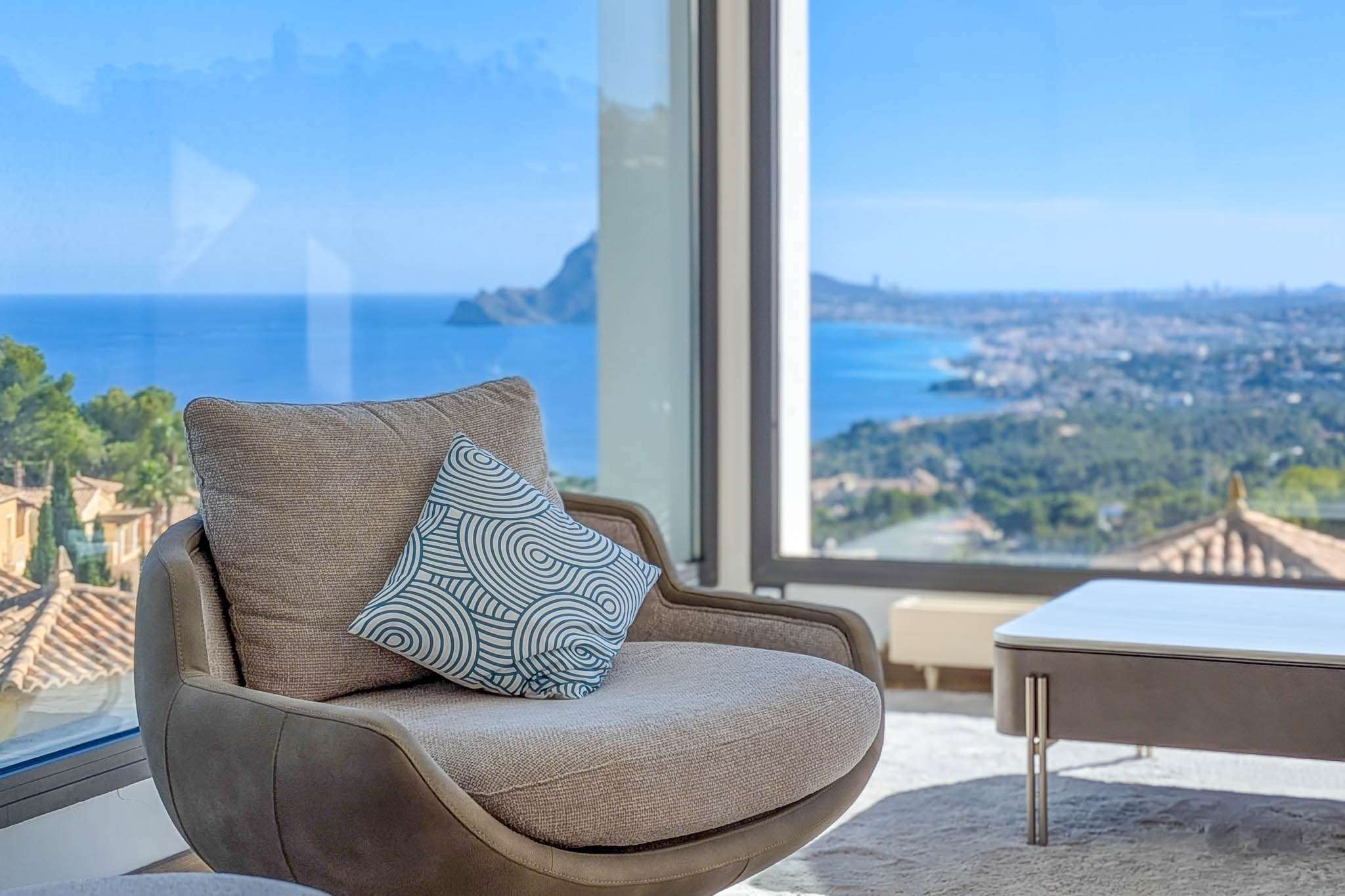 Gemütlicher Sessel am Panoramafenster mit unverbautem Blick auf das Mittelmeer und die Küstenlinie. Ein Zierkissen ergänzt das moderne Interieurdesign.