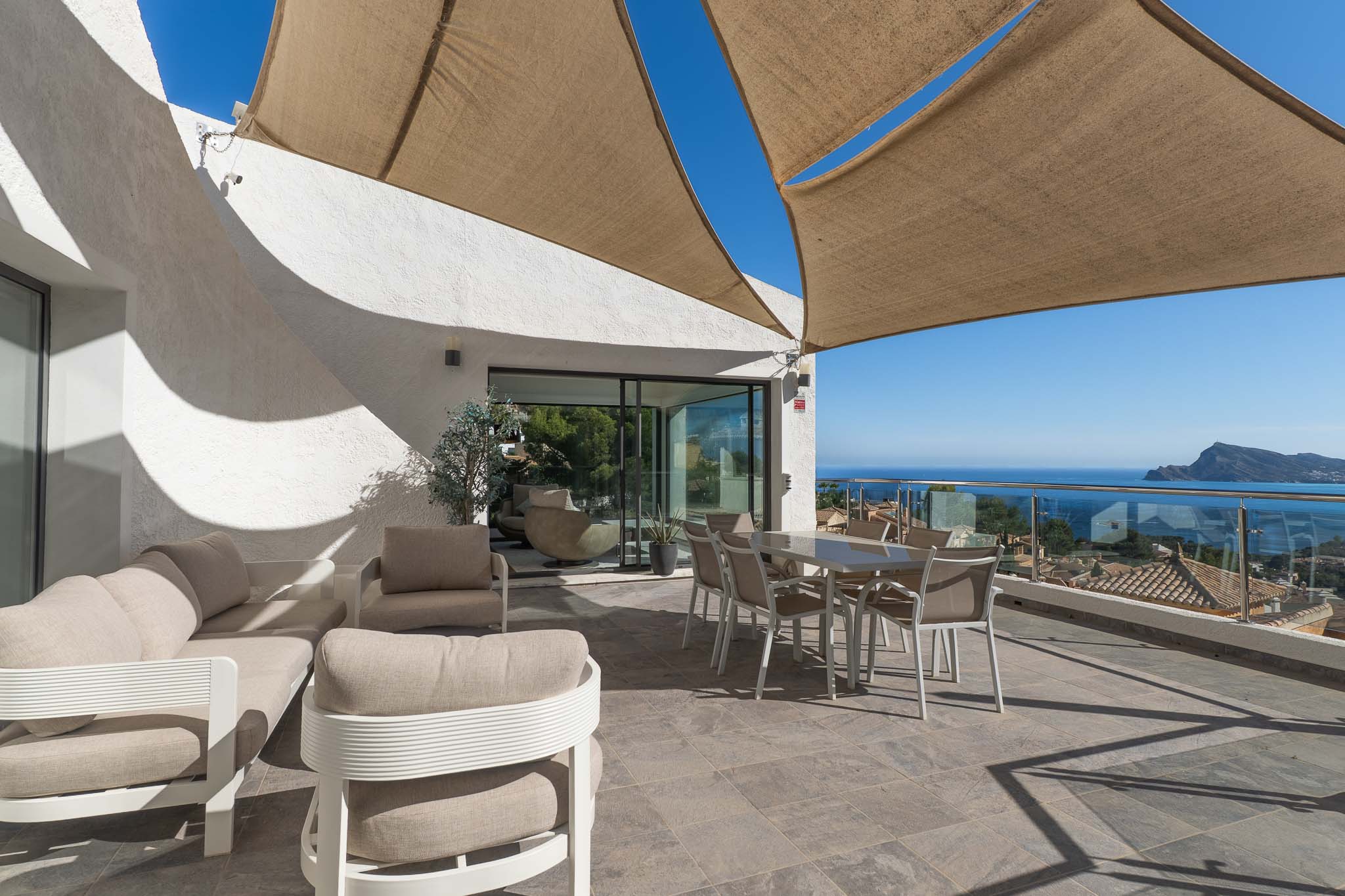 Geräumige Außenterrasse mit Lounge- und Essbereichen unter einem Sonnensegel, mit Panoramablick auf Meer und Küste durch eine Glasbrüstung.