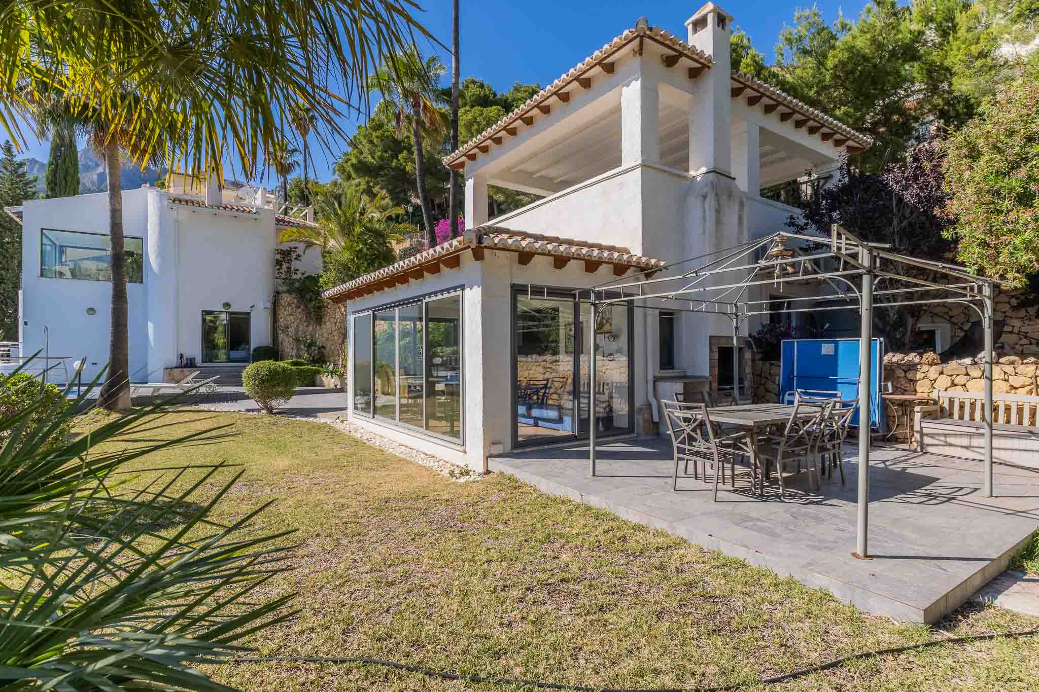 Geräumige gepflasterte Terrasse mit Essbereich unter einer Metallpergola, angrenzend an einen verglasten Raum und einen gepflegten mediterranen Garten.