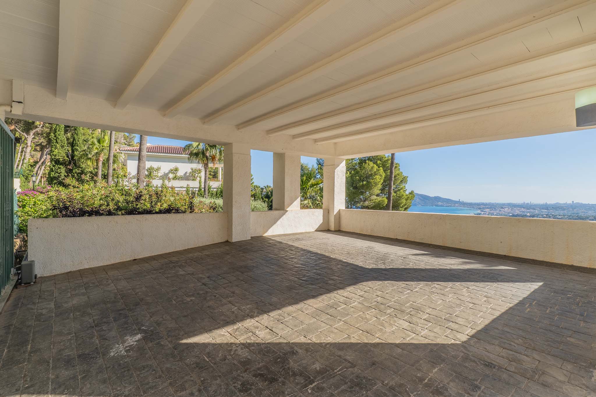 Geräumiger überdachter Carport mit gestanztem Betonboden und weißer Balkendecke. Er bietet Panoramablicke auf das Mittelmeer und die Küste, umgeben von üppiger Vegetation.