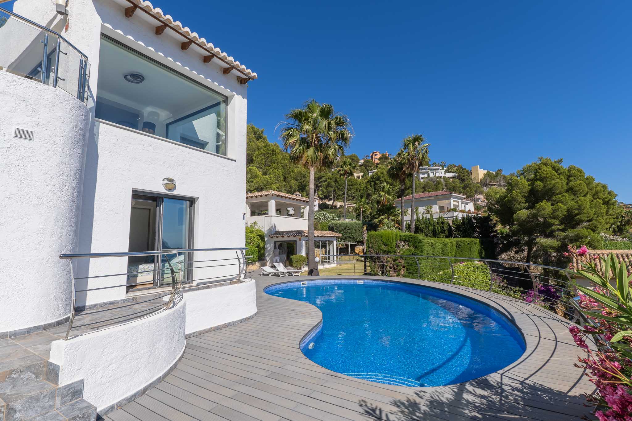 Geschwungener blauer Mosaikpool mit grauer Verbundterrasse, angrenzend an eine weiße Fassade mit Panoramafenster und Balkon mit Edelstahlgeländer.