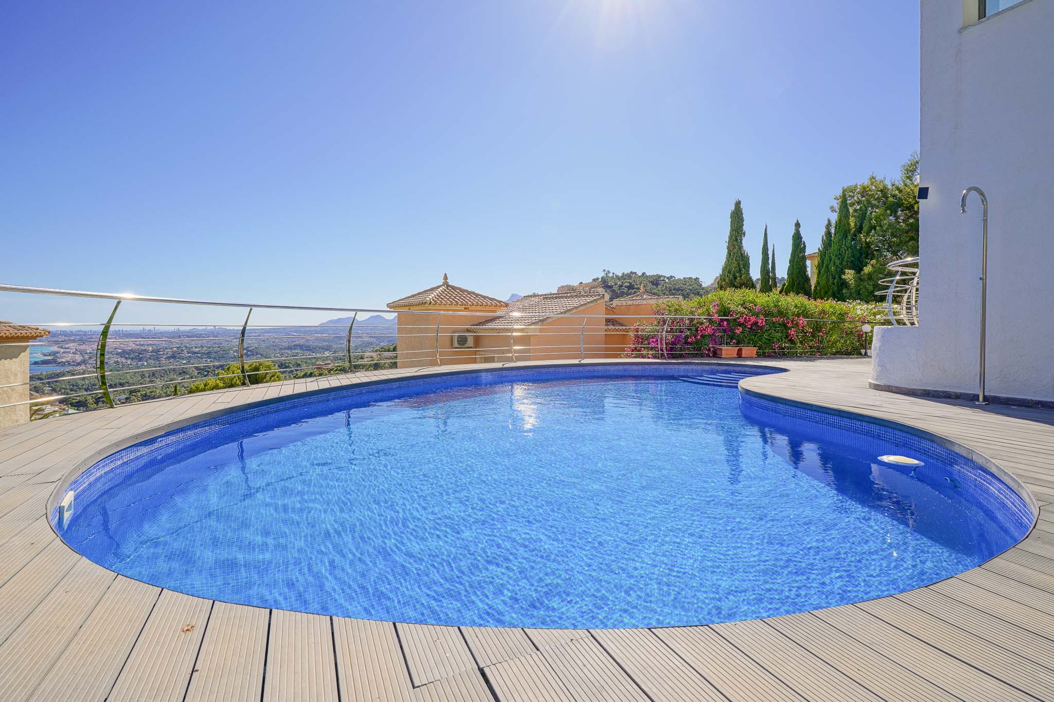 Geschwungener Pool mit blauen Mosaikfliesen und heller Holzterrasse, umgeben von einem Edelstahlgeländer, mit Panoramablick auf Tal und ferne Stadt.