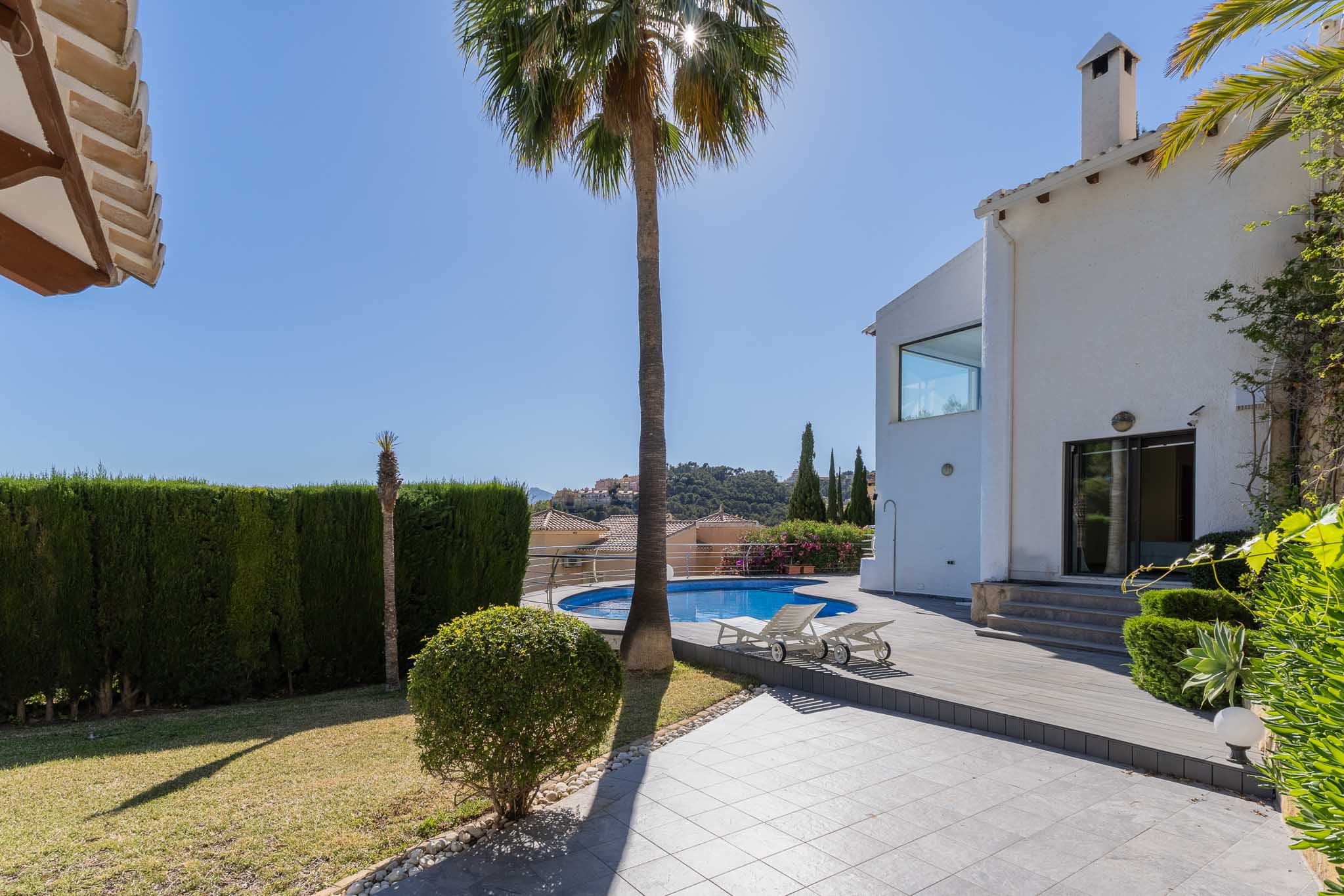 Großer Garten mit rundem Pool und Holzdeck-Solarium, begrenzt von einer weißen Villa und einer hohen Hecke. Blick auf die Hügellandschaft.