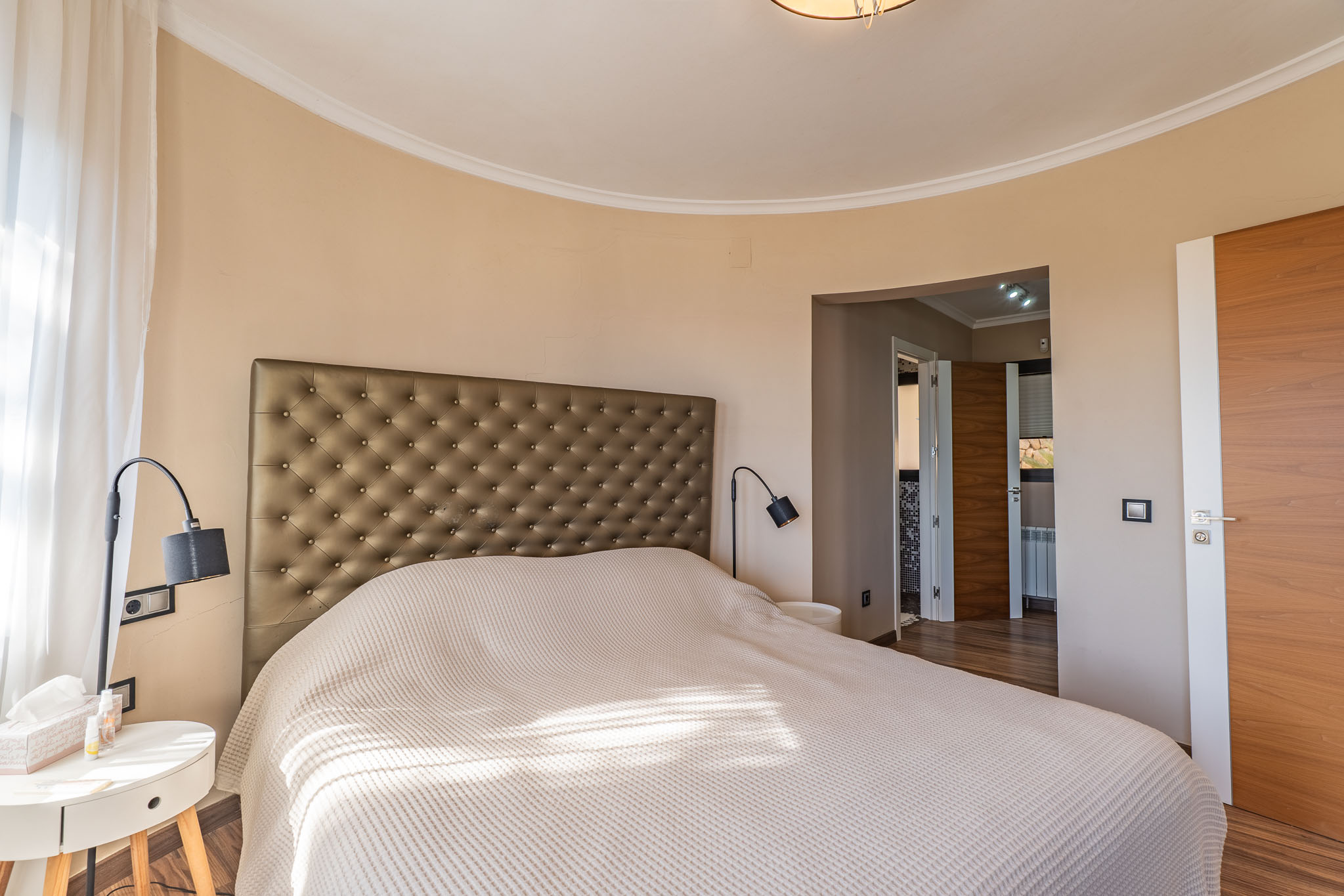 Hauptschlafzimmer mit gepolstertem Kopfteil, einer markanten geschwungenen Wand und natürlichem Licht durch transparente Vorhänge, ergänzt durch Holztüren.