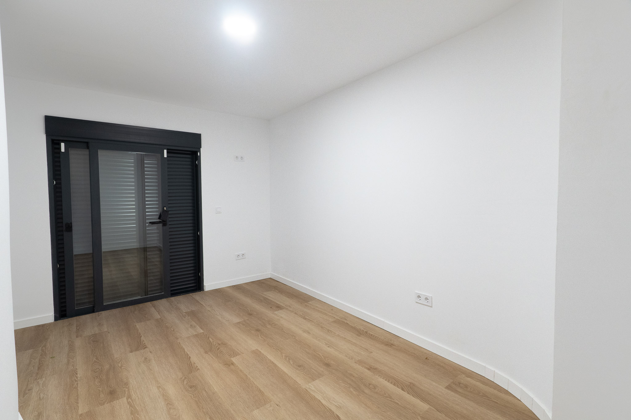 Heller Raum mit hellem Holzlaminatboden und weißen Wänden. Eine große anthrazitfarbene Glasschiebetür mit integrierten Jalousien bietet Zugang zum Außenbereich.