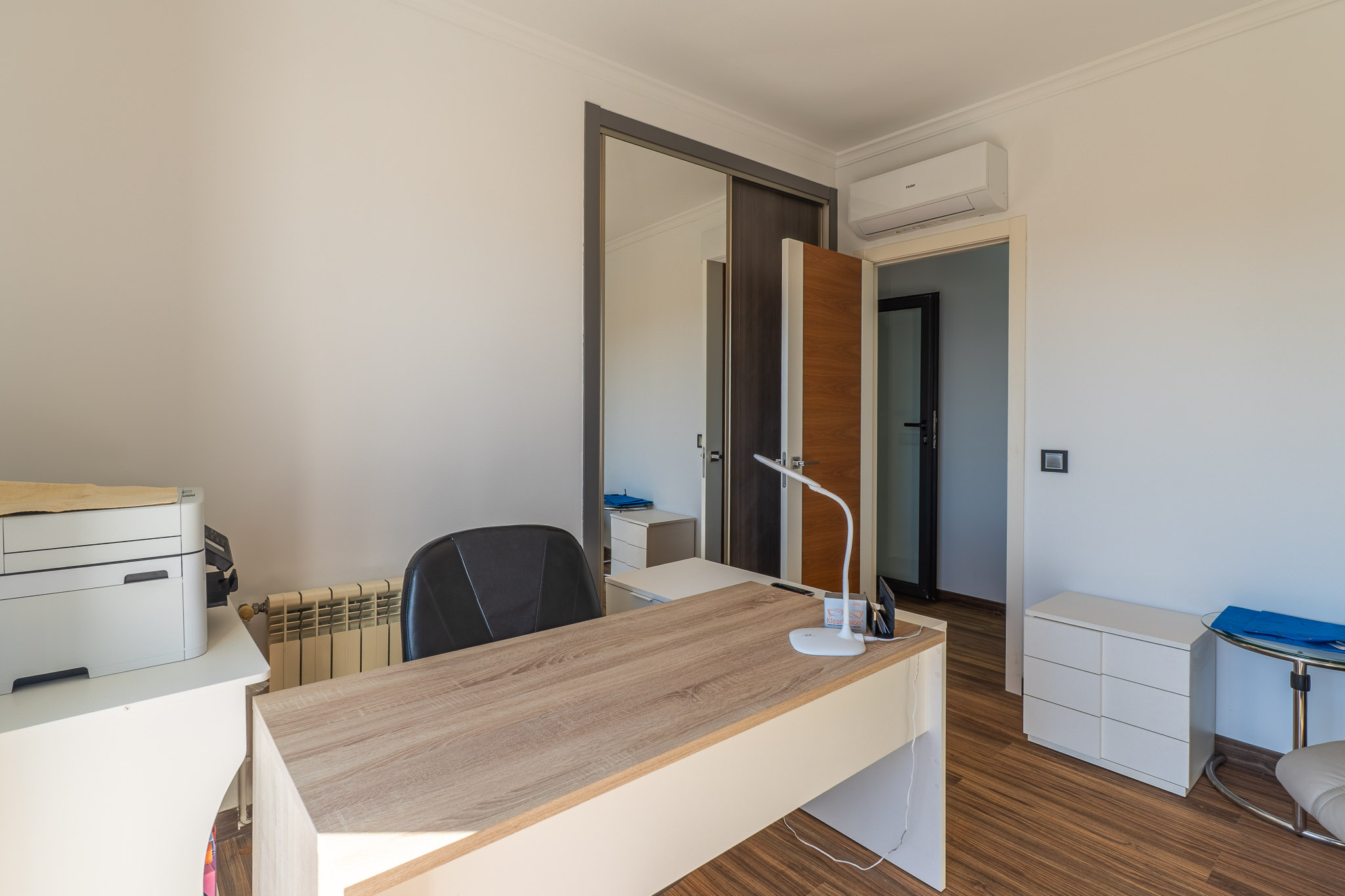Helles Arbeitszimmer mit hellem Holzschreibtisch, Laminatboden, verspiegeltem Schiebetürenschrank und Klimaanlage.
