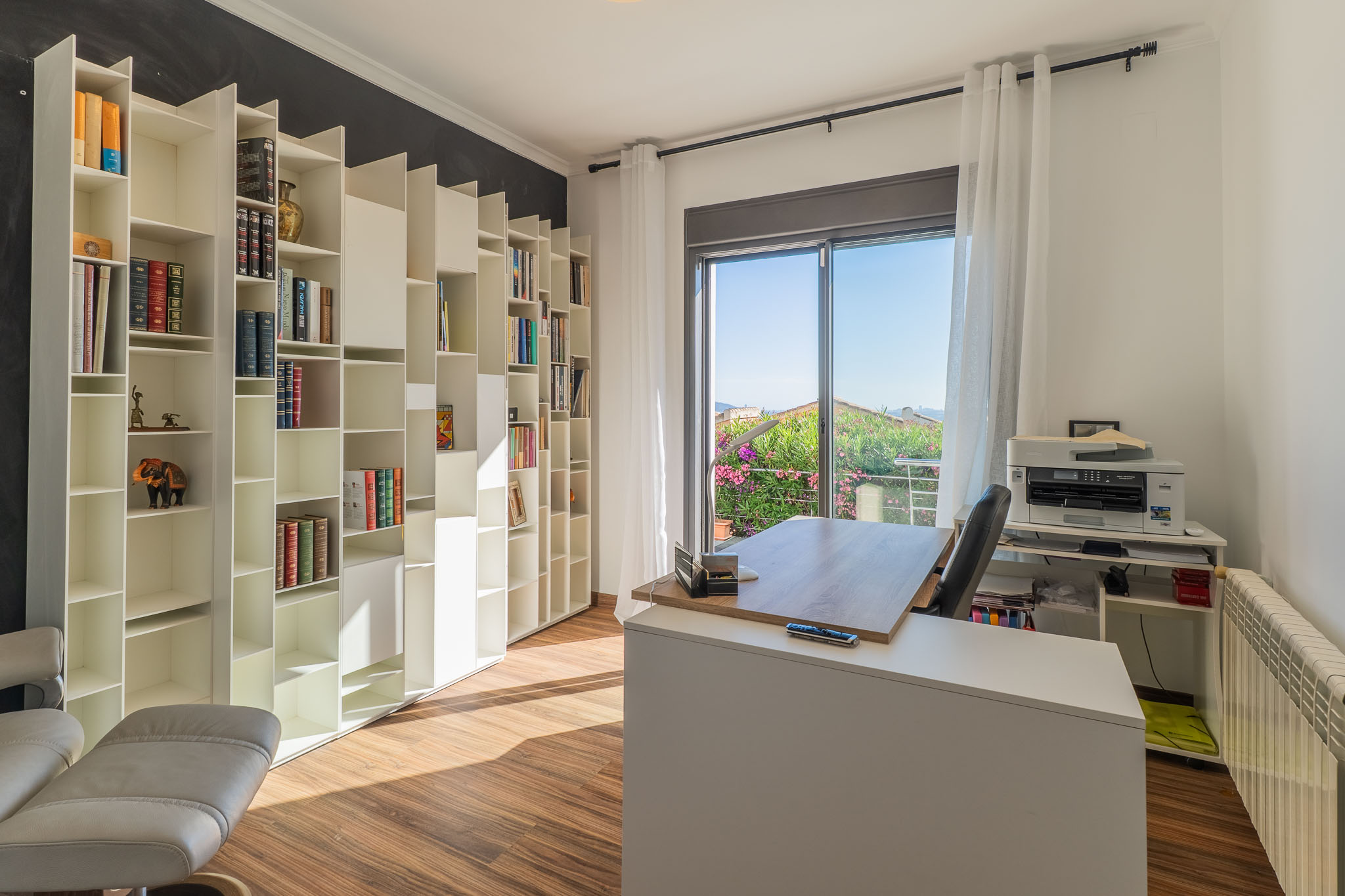 Helles Arbeitszimmer mit weißem modularem Bücherregal, Holzboden und modernem Schreibtisch vor einem Balkon mit üppiger Gartenansicht. Natürliches Licht durchflutet den Raum.