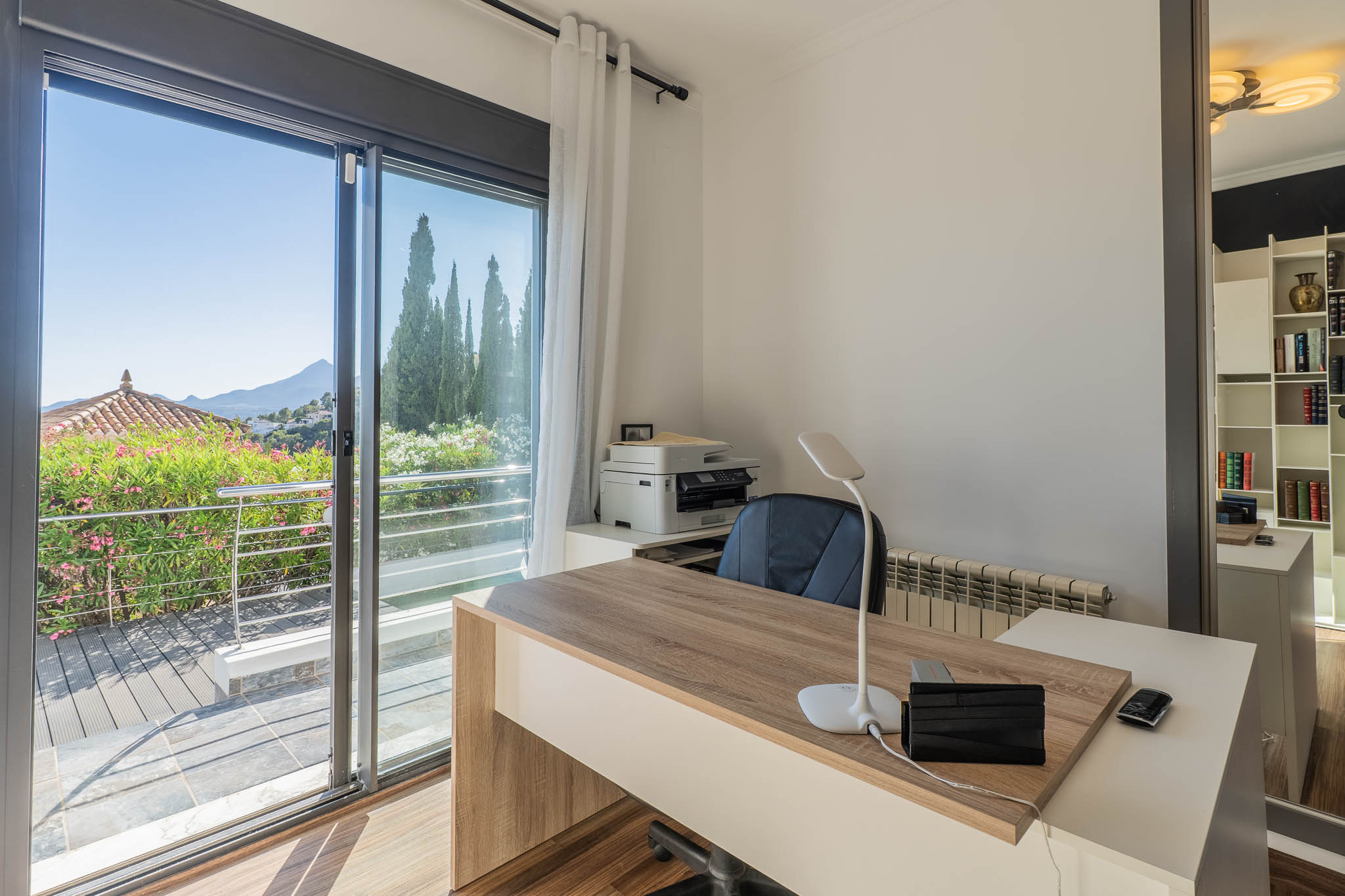 Helles Büro mit modernem Holzschreibtisch, großer Glasschiebetür zu einer Terrasse mit üppigem Grün und Panoramablick auf die Berge. Ein Spiegel reflektiert ein praktisches Bücherregal.