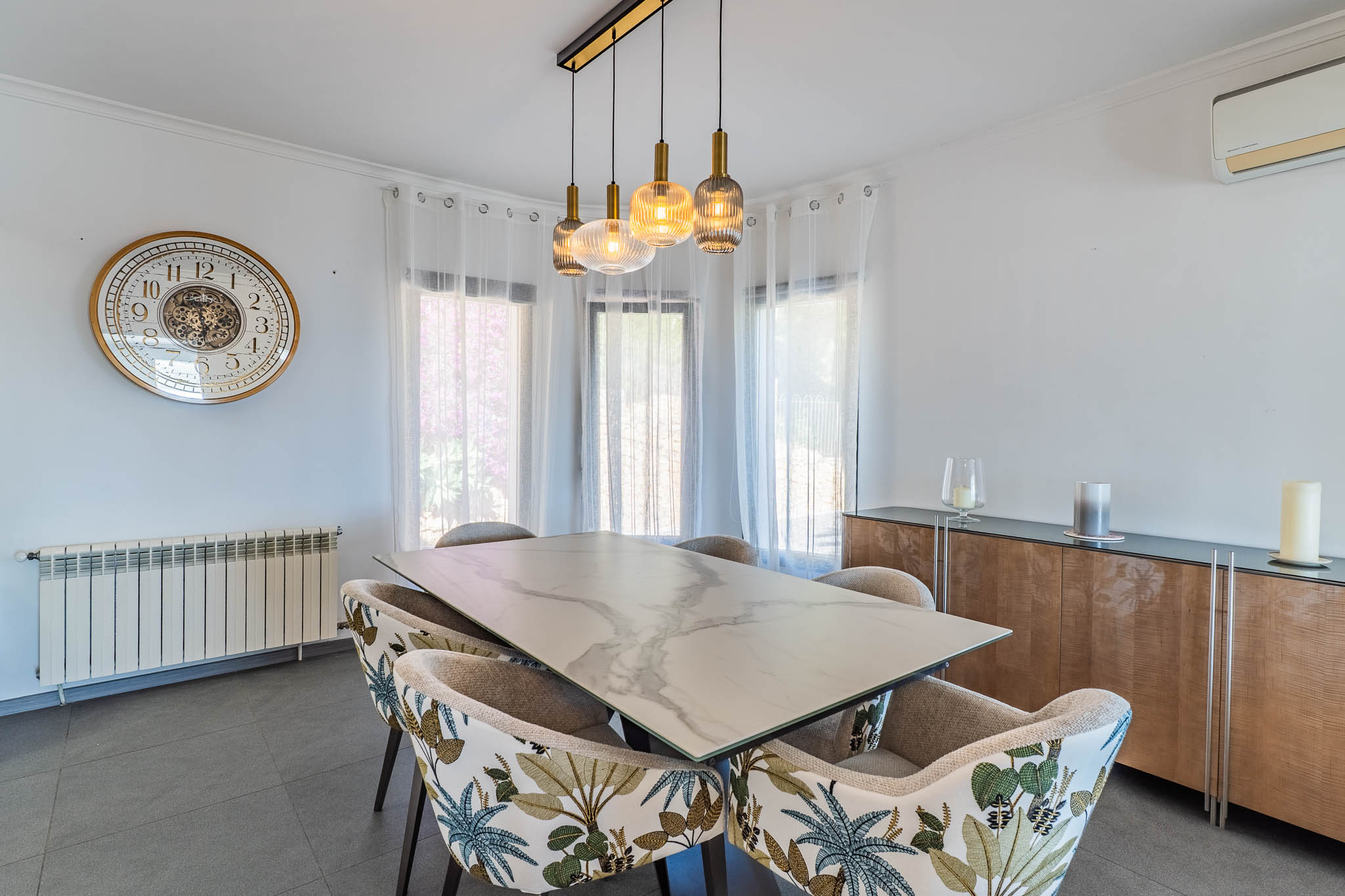 Helles Esszimmer mit Marmoreffekt-Tisch, Stühlen mit tropischem Muster und einer modernen linearen Hängeleuchte, von Tageslicht erhellt.