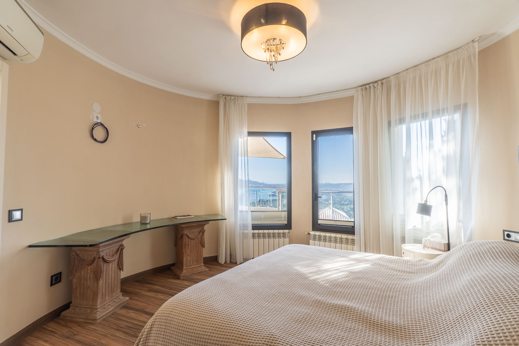 Helles Schlafzimmer mit Panoramameerblick durch große Fenster mit Vorhängen und einem einzigartigen Glastisch auf geschnitzten Holzsockeln.