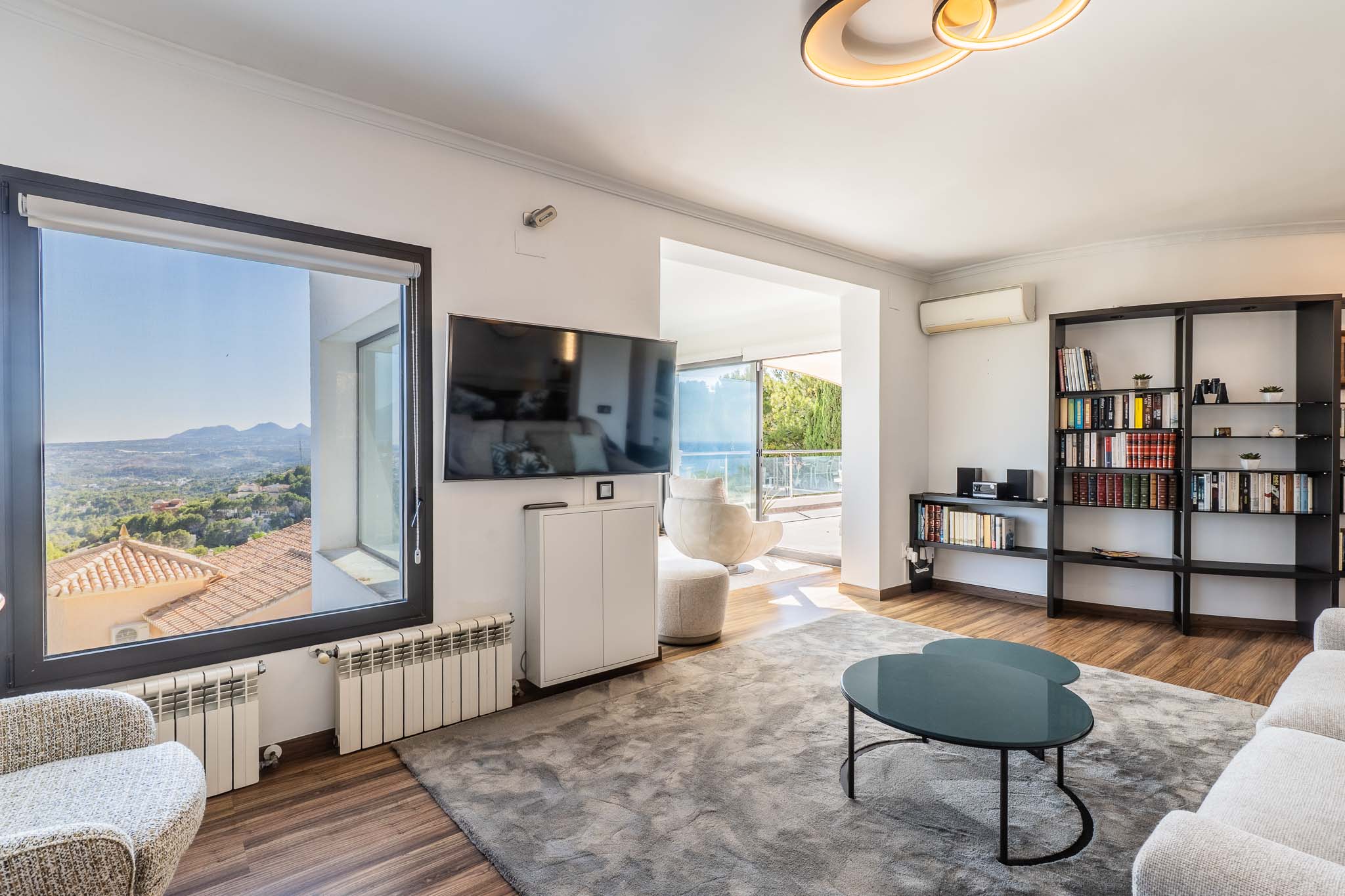 Helles Wohnzimmer mit Panoramablick auf die Berge vom Fenster und Zugang zur Terrasse mit Meerblick. Ausgestattet mit Holzboden, einem großen grauen Teppich und einem modernen Bücherregal.