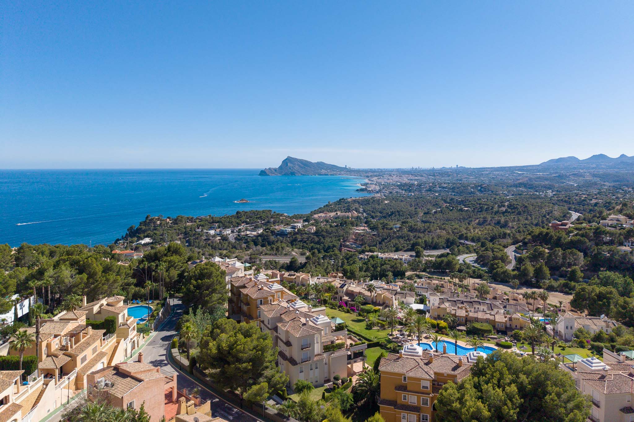 Luftaufnahme der Küste von Altea Hills, mit dem Mittelmeer, dem Peñón de Ifach und der Skyline von Benidorm in der Ferne. Terrakottadächer und Pools sind inmitten üppiger Vegetation sichtbar.