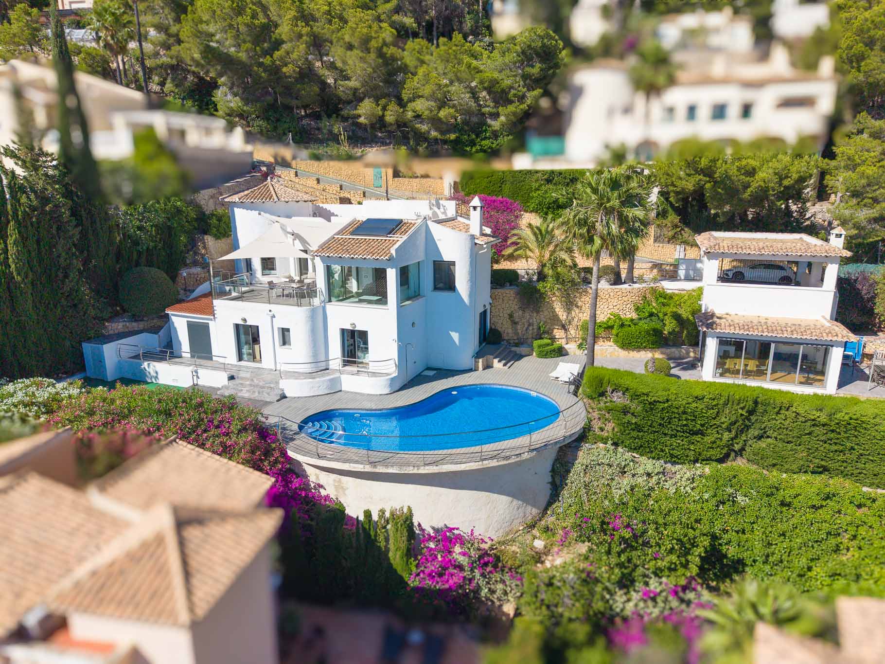 Luftaufnahme der weißen Villa, mit einem einzigartigen nierenförmigen Pool, umgeben von einer Holzterrasse und Metallgeländer. Mehrere Terrassen und üppige mediterrane Gärten sind sichtbar.