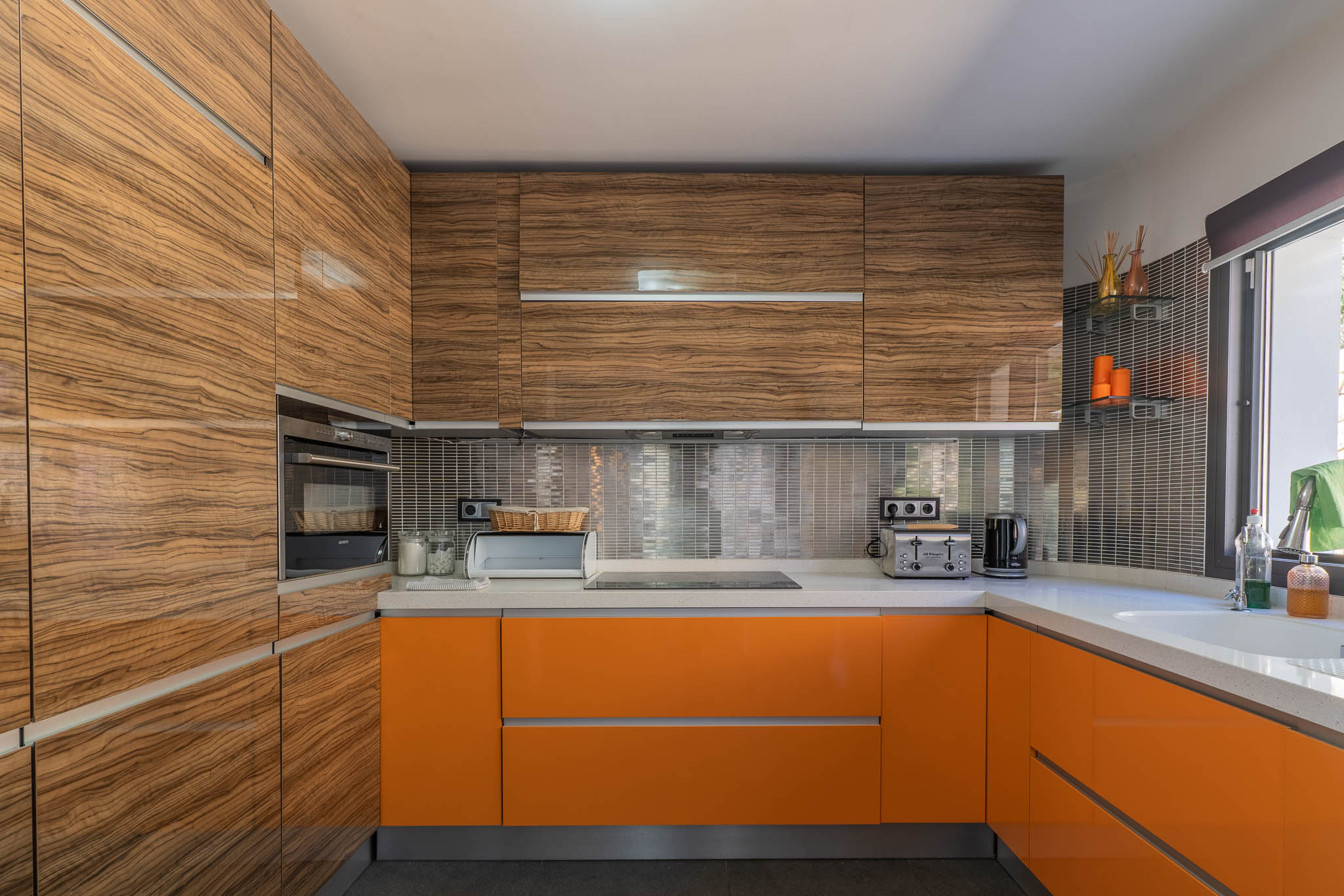 Moderne Küche mit glänzenden Holzmaser-Oberschränken und leuchtend orangefarbenen Unterschränken, ergänzt durch helle Arbeitsplatten und einen metallischen Mosaik-Spritzschutz.