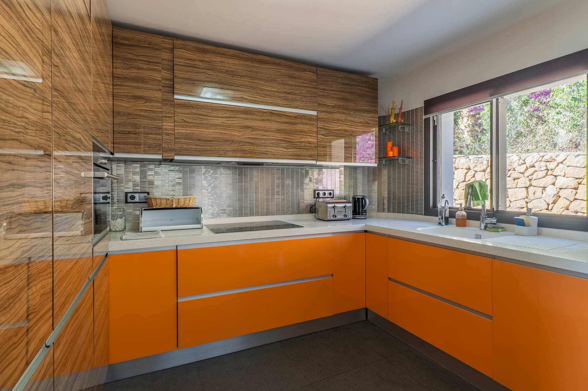 Moderne Küche mit glänzenden Holzmaser-Oberschränken und leuchtend orangefarbenen Unterschränken. Weiße Arbeitsplatte, metallische Mosaik-Rückwand und ein Fenster mit Blick auf eine Steinmauer.