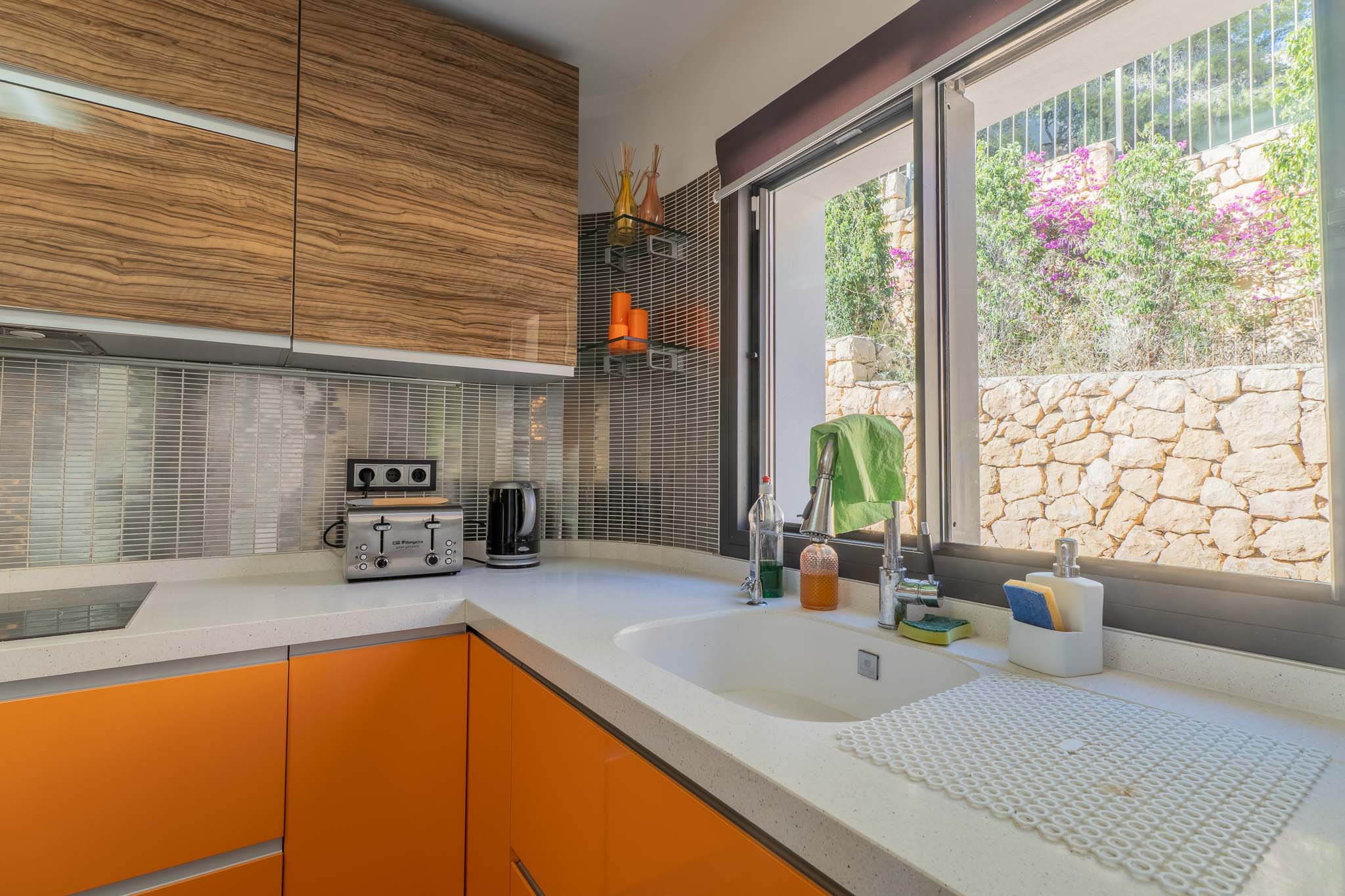 Moderne Küche mit weißer Quarz-Arbeitsplatte, leuchtend orangefarbenen Unterschränken und Oberkästen in Holzoptik. Das Fenster blickt auf eine Natursteinmauer und Pflanzen.