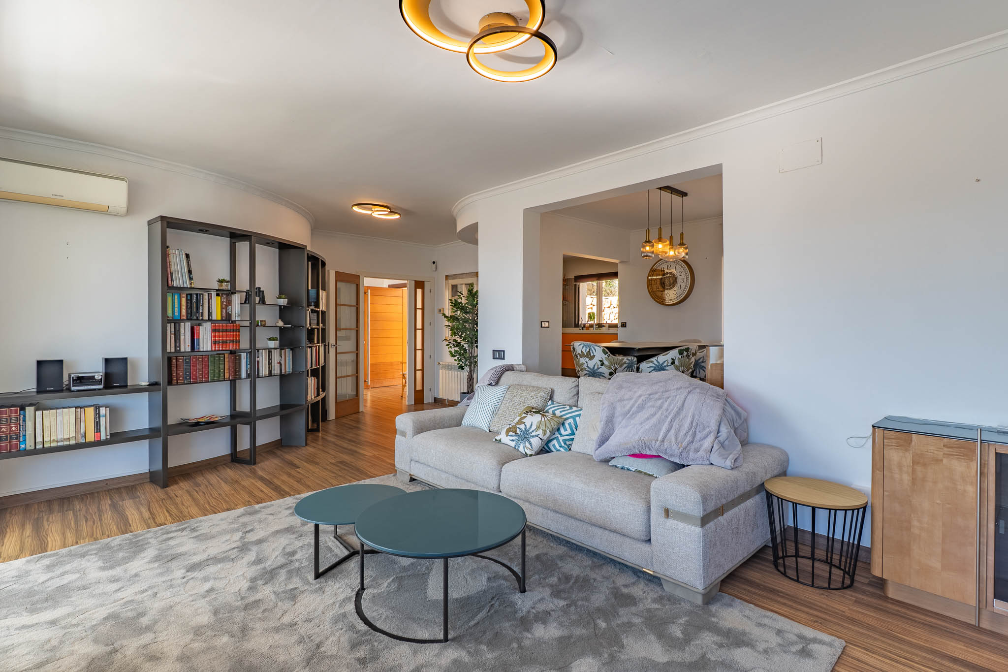 Moderner Wohnbereich mit hellem Sofa, zwei runden Teetischen in Petrol und einem großen modularen Bücherregal. Design-Deckenleuchten erhellen den Raum.
