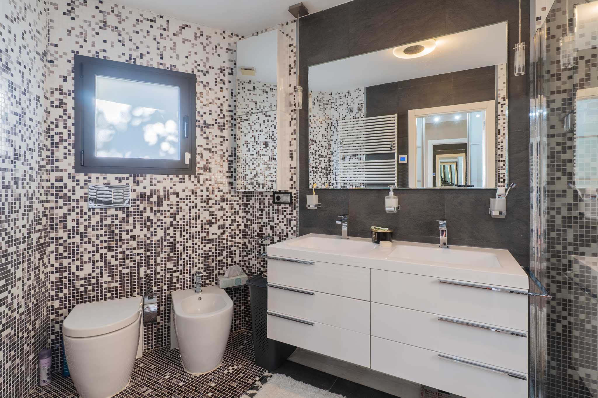 Modernes Bad mit Mosaikfliesen an Wänden und Boden in Erd-/Grautönen. Es bietet einen schwebenden Doppelwaschtisch, großen beleuchteten Spiegel, wandhängendes WC, Bidet und Handtuchwärmer.