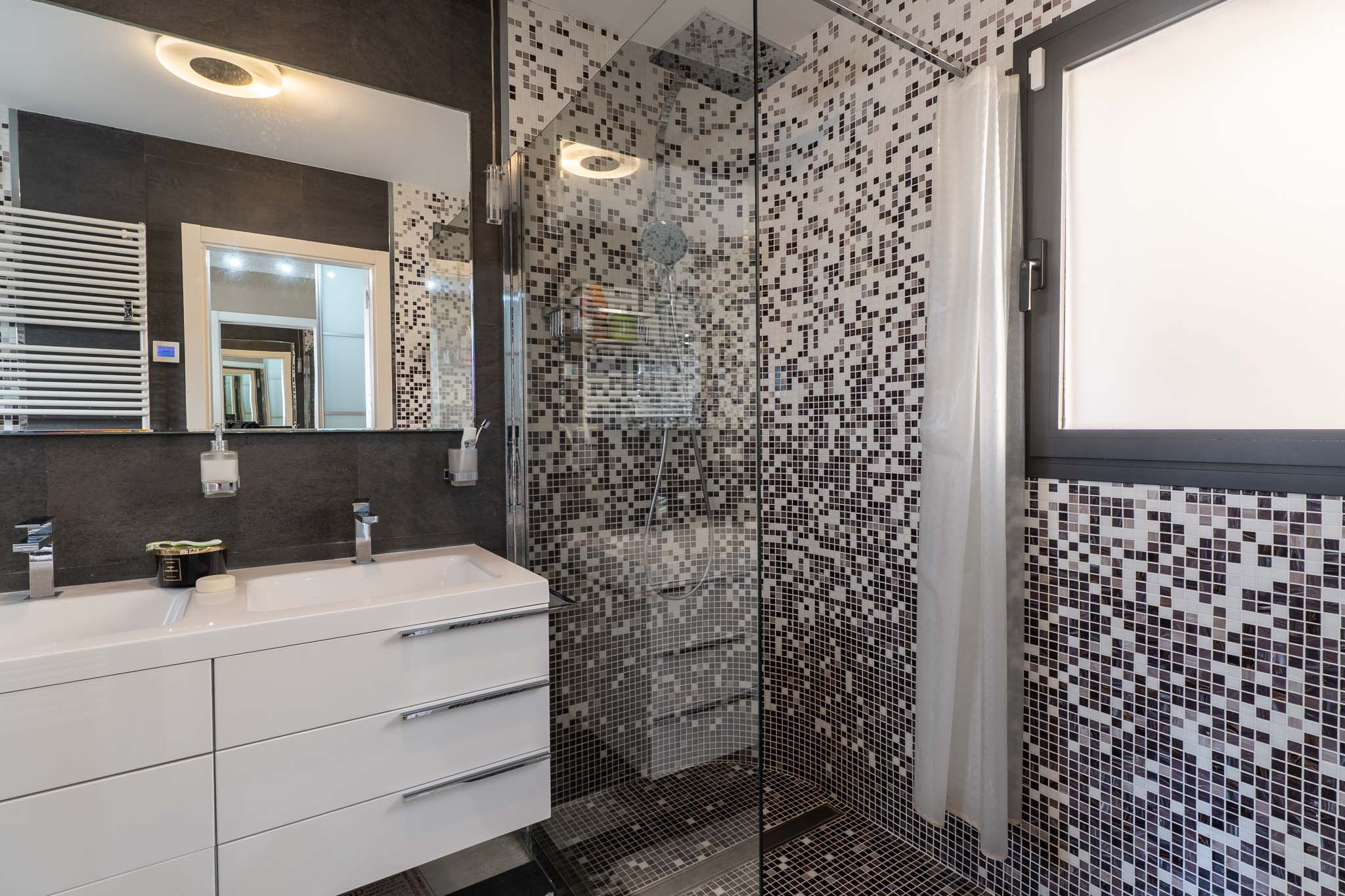 Modernes Badezimmer mit Doppelwaschtisch, weißen Unterschränken und beleuchtetem Spiegel. Die Glasdusche hat Mosaikfliesen in Grautönen und ein integriertes Fenster.