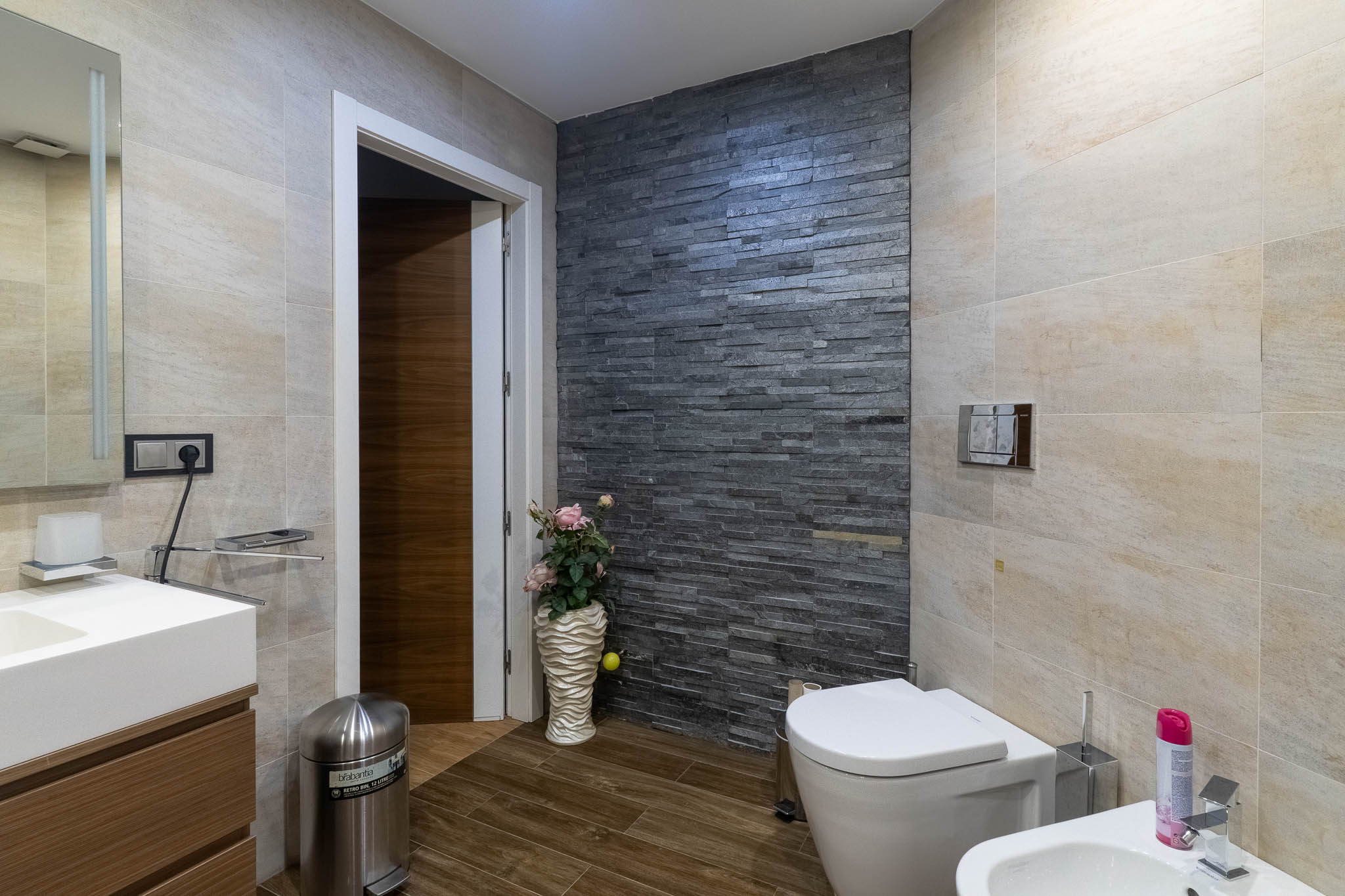 Modernes Badezimmer mit dunkler Natursteinwand, hellbeigen Fliesen und Holzoptikboden. Ausgestattet mit WC, Bidet und Waschtisch mit Waschbecken.