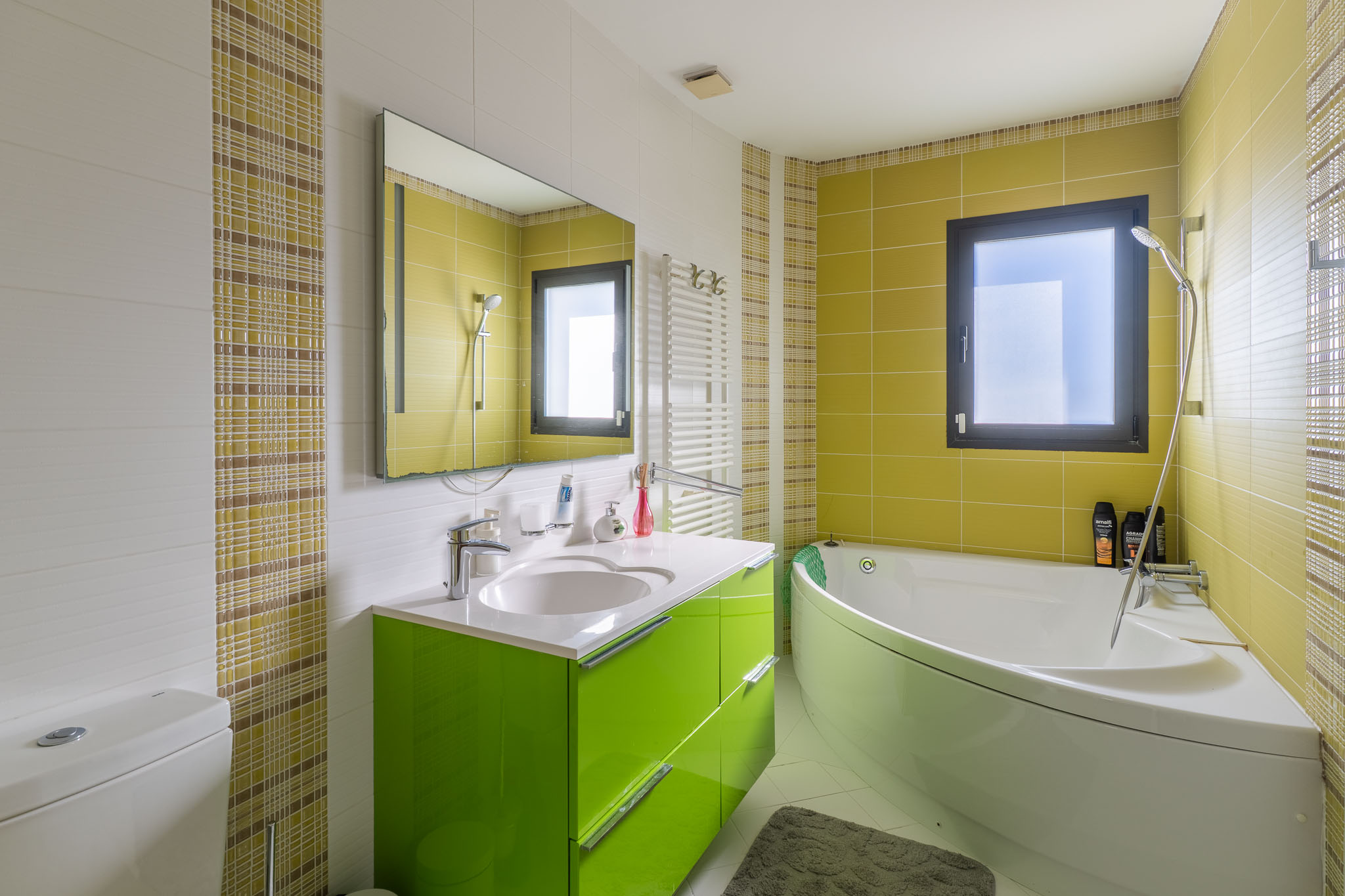Modernes Badezimmer mit Eckbadewanne und leuchtend grünem Waschtisch. Wände gefliest mit weißen Struktur-, lindgrünen und karierten Mosaikfliesen. Mit Fenster und Handtuchheizkörper.