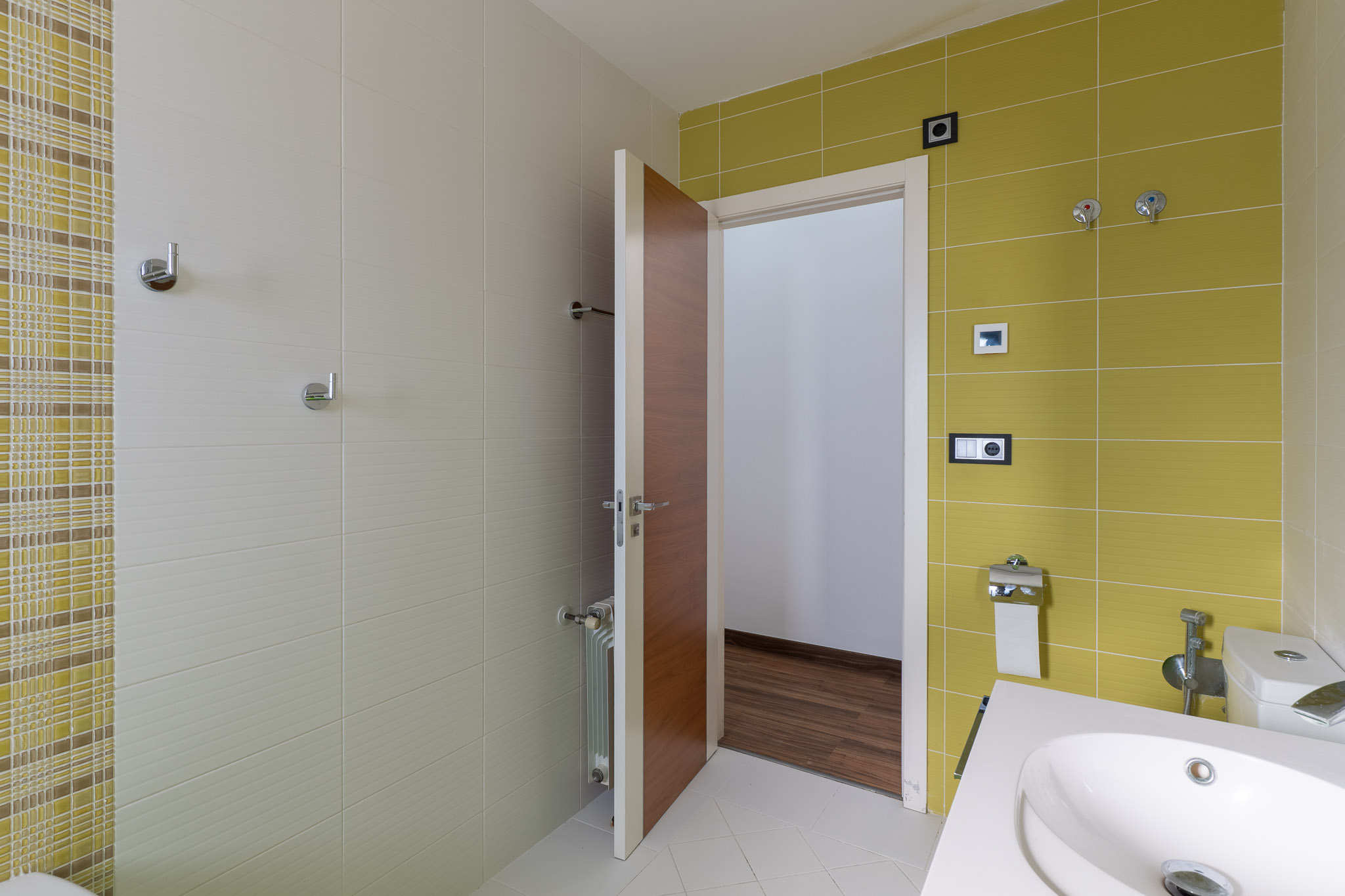 Modernes Badezimmer mit Wandfliesen in Limettengrün und strukturiertem Beige, ergänzt durch ein Mosaikband. Ausgestattet mit einem zeitgemäßen Waschbecken und einer Holztür.
