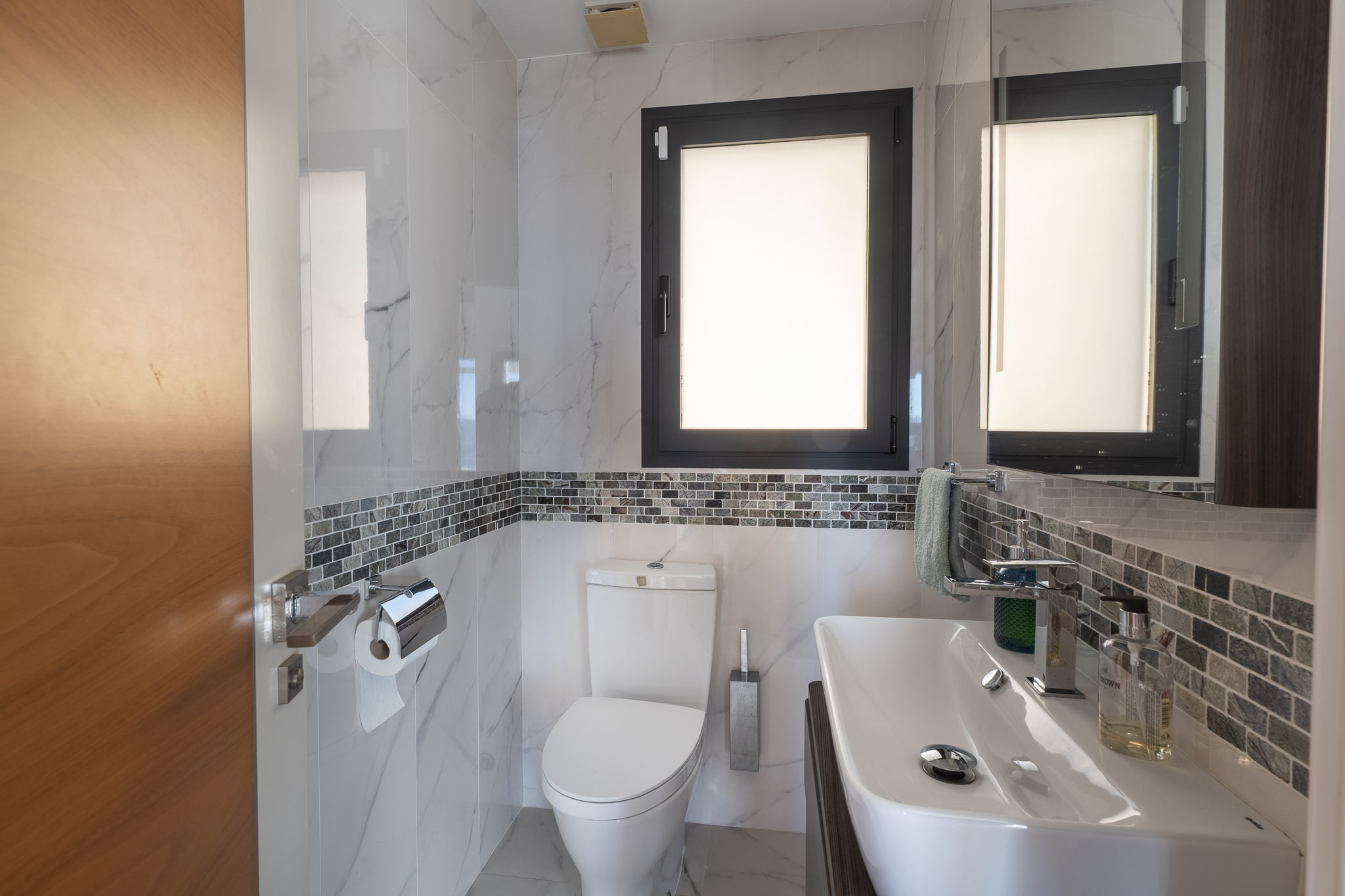 Modernes Badezimmer mit weißen Marmor-Effektfliesen, einem grauen Mosaikband und einem dunkel gerahmten Fenster. Mit elegantem weißen Waschbecken und passender Toilette.