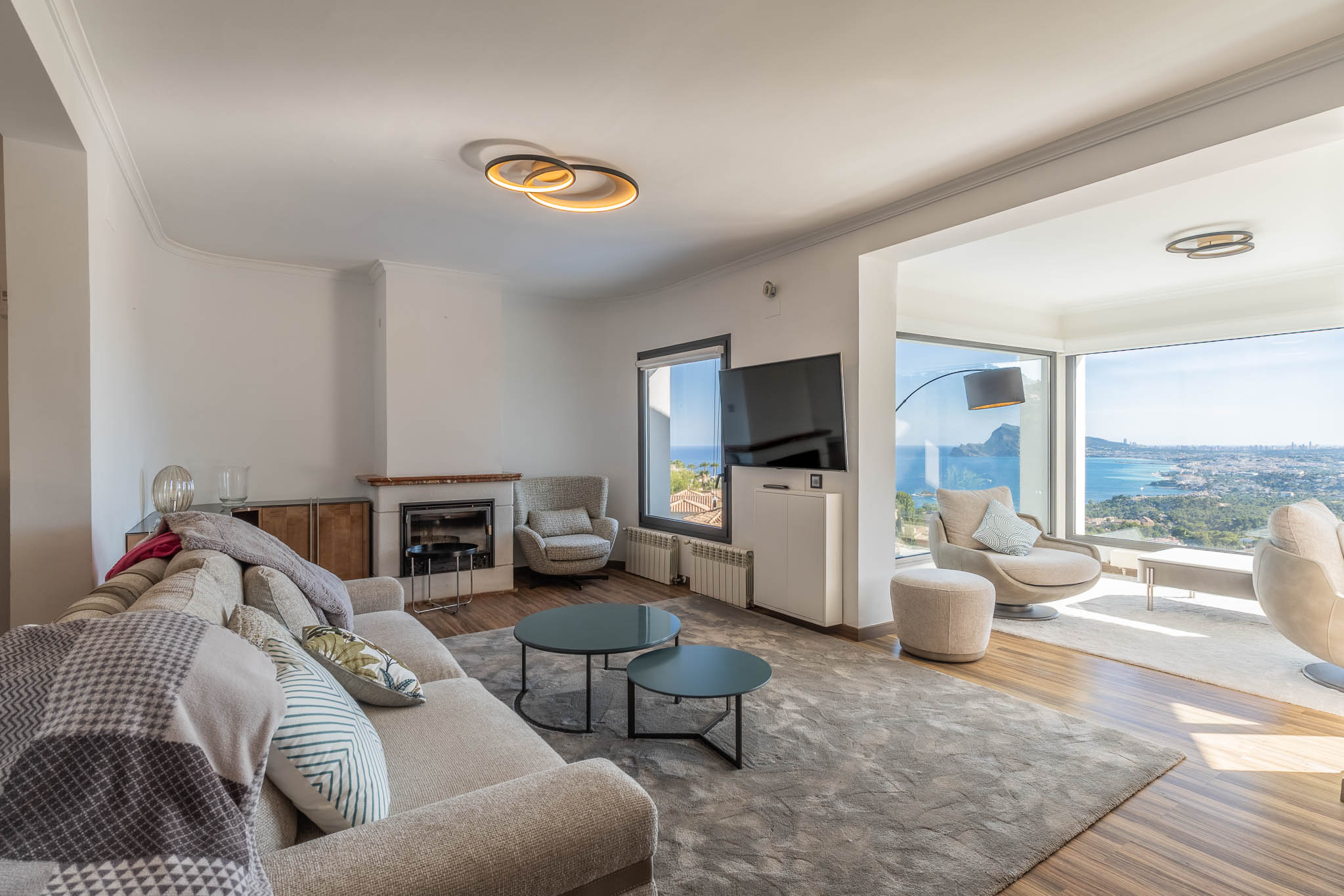Modernes Wohnzimmer mit hellem Sofa, Kamin und großen Fenstern, die einen Panoramablick auf Meer und Stadt bieten. Die zeitgenössische Einrichtung hellt den Raum auf.