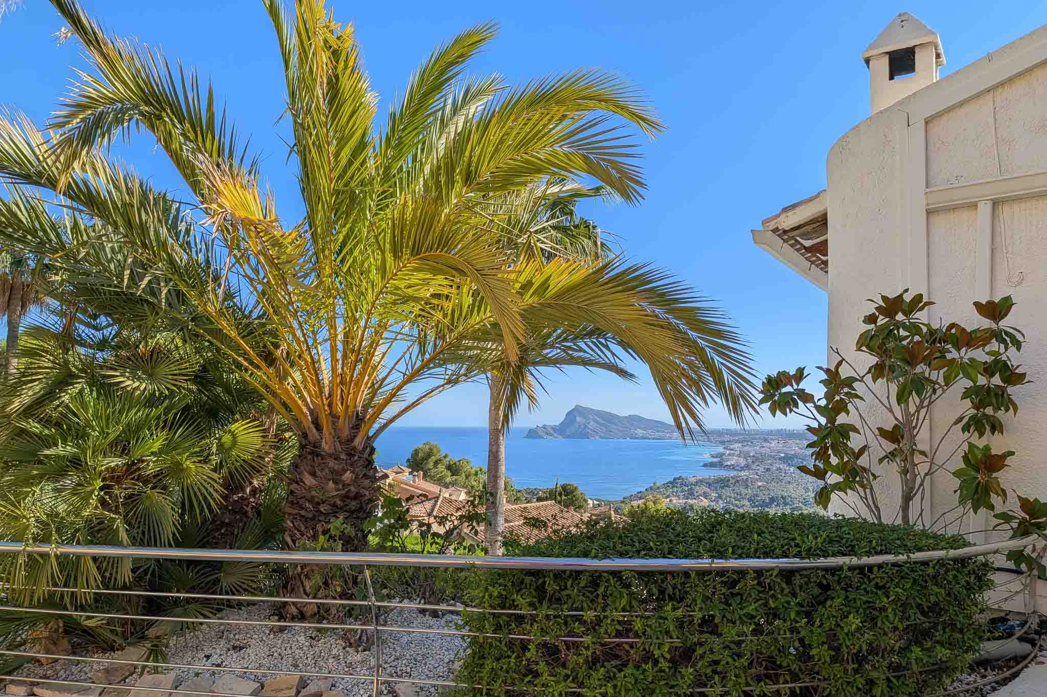 Panoramablick auf das Meer von einer Terrasse, umrahmt von üppigen Palmen. Edelstahlgeländer, niedrige Hecke und die Küste mit dem ikonischen Peñón de Ifach.