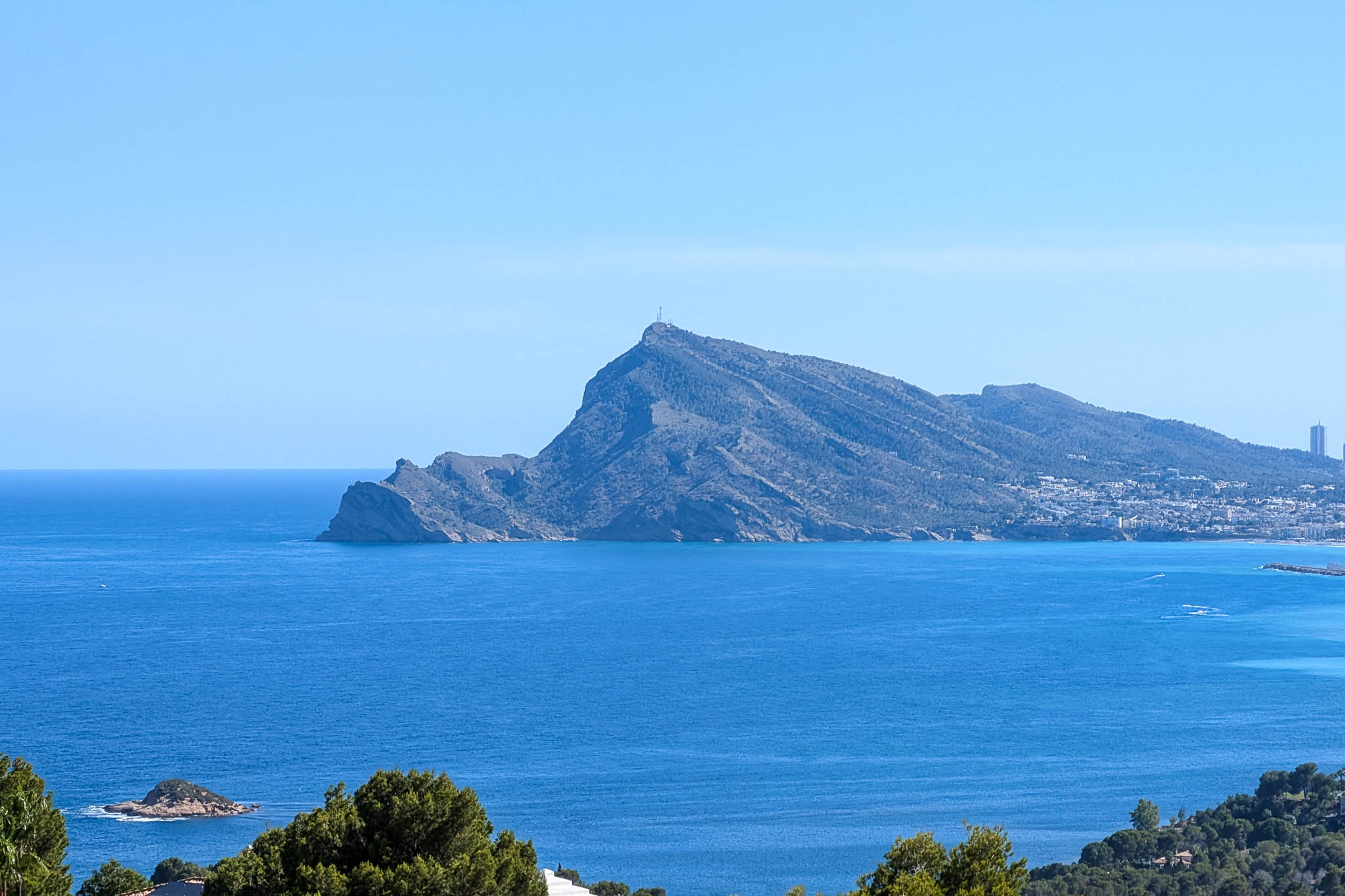 Panoramablick auf das Mittelmeer mit dem markanten Profil der Sierra Helada, einer entfernten Küstenstadt und kleinen Felseninseln unter klarem blauem Himmel.