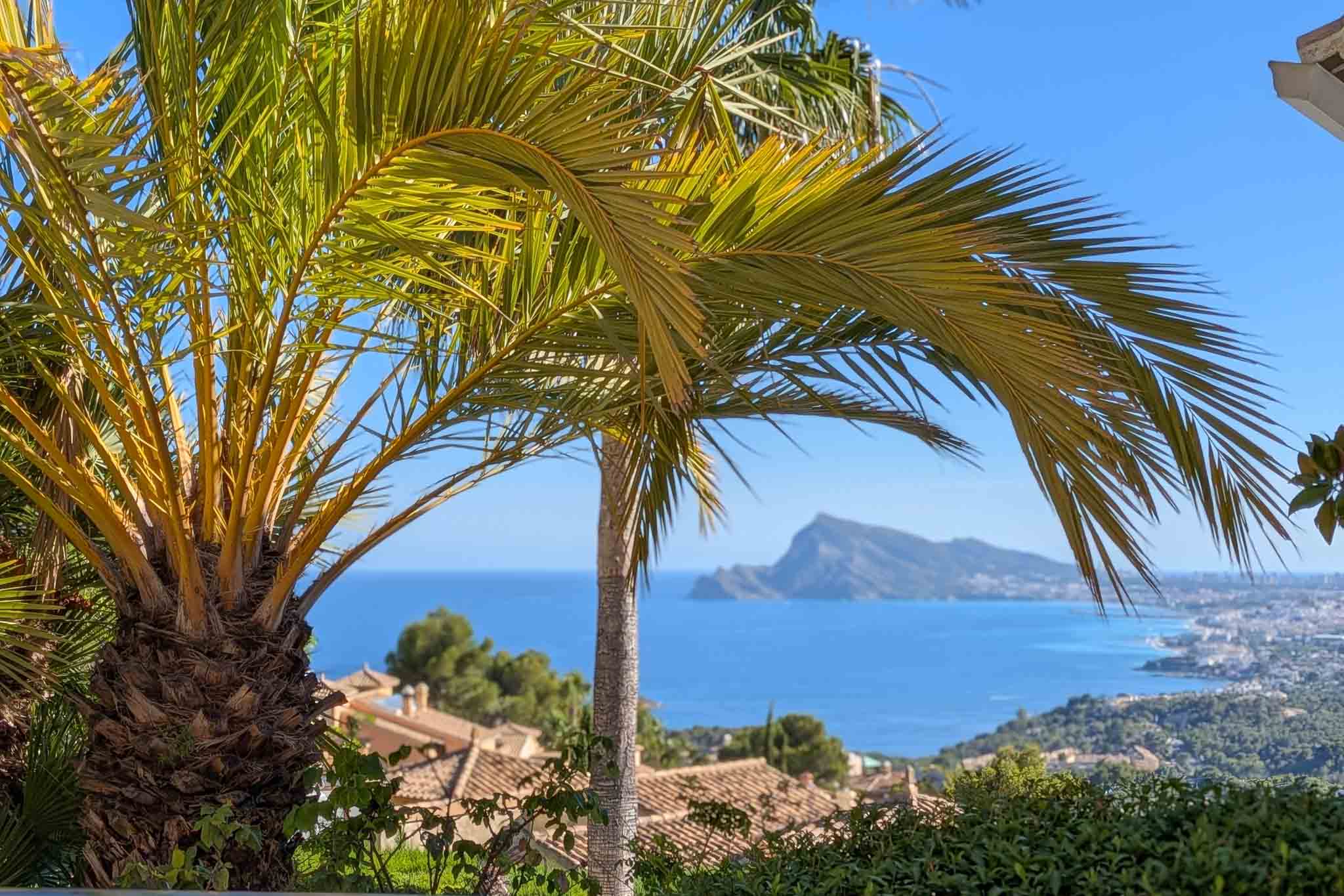 Panoramablick auf das Mittelmeer, umrahmt von Palmen, mit den Dächern von Altea Hills und dem fernen Peñón de Ifach unter blauem Himmel.