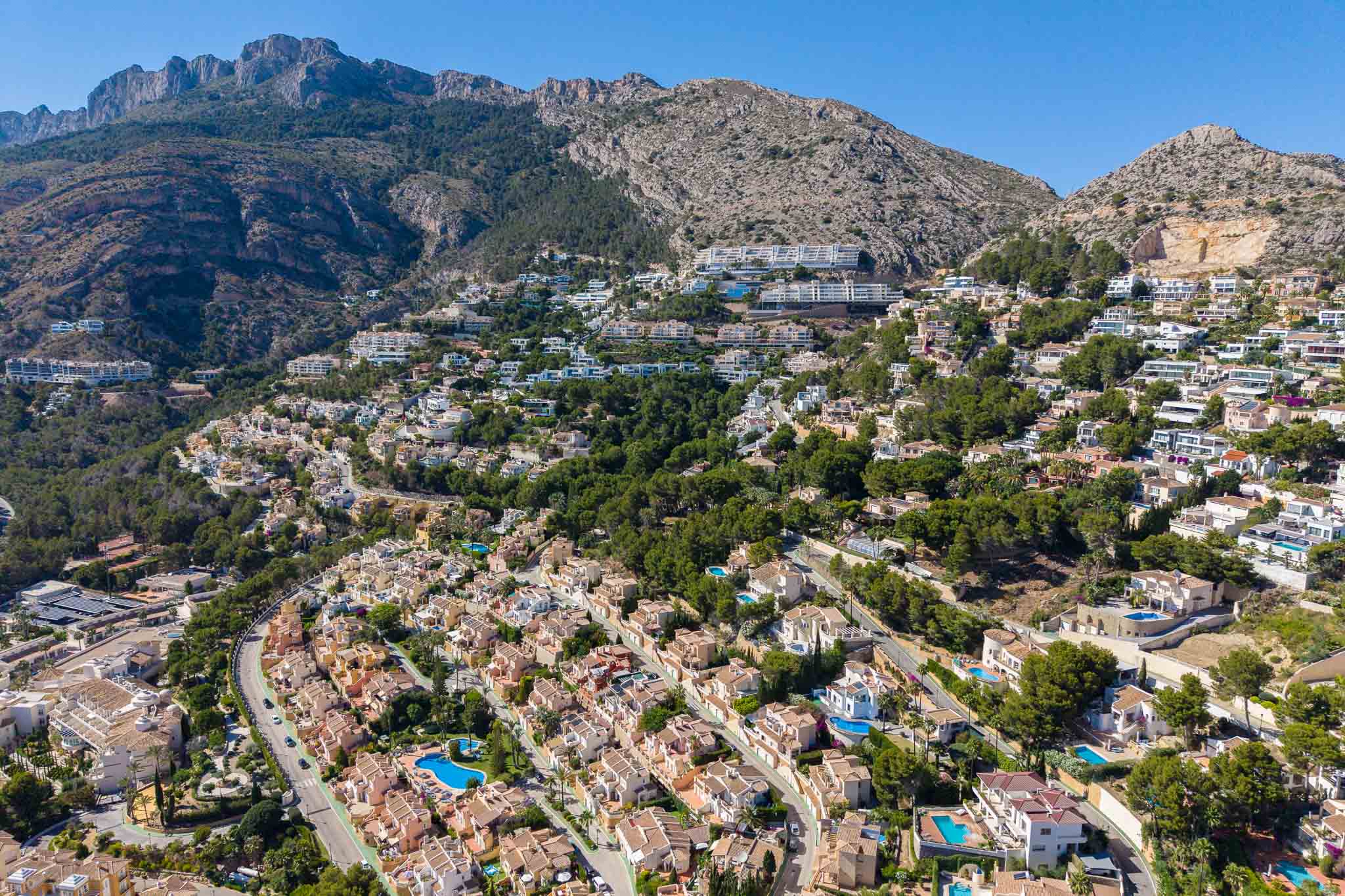 Panoramische Luftaufnahme von Altea Hills, die zahlreiche Villen mit Pools an einem grünen Hang vor einer dramatischen Bergkulisse zeigt.