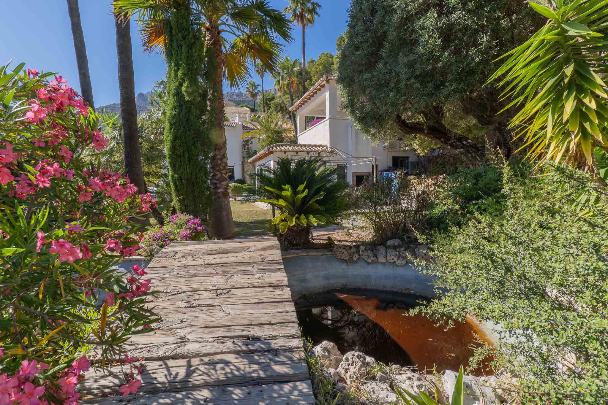 Rustikaler Holzsteg über einem ruhigen Gartenteich, umrahmt von leuchtend rosa Oleander und hohen Palmen, die zur Villa unter mediterraner Sonne führen.