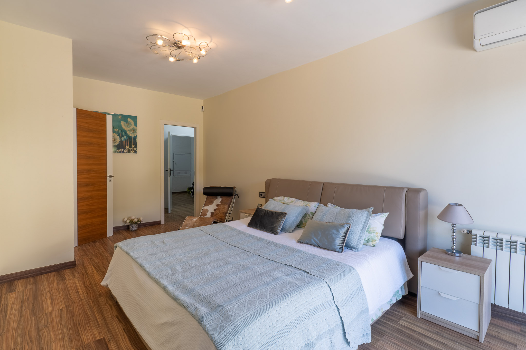 Schlafzimmer mit beigem Kopfteilbett, Laminatboden und moderner Deckenleuchte. Nachttisch mit Lampe, Heizkörper und Klimaanlage sind sichtbar.
