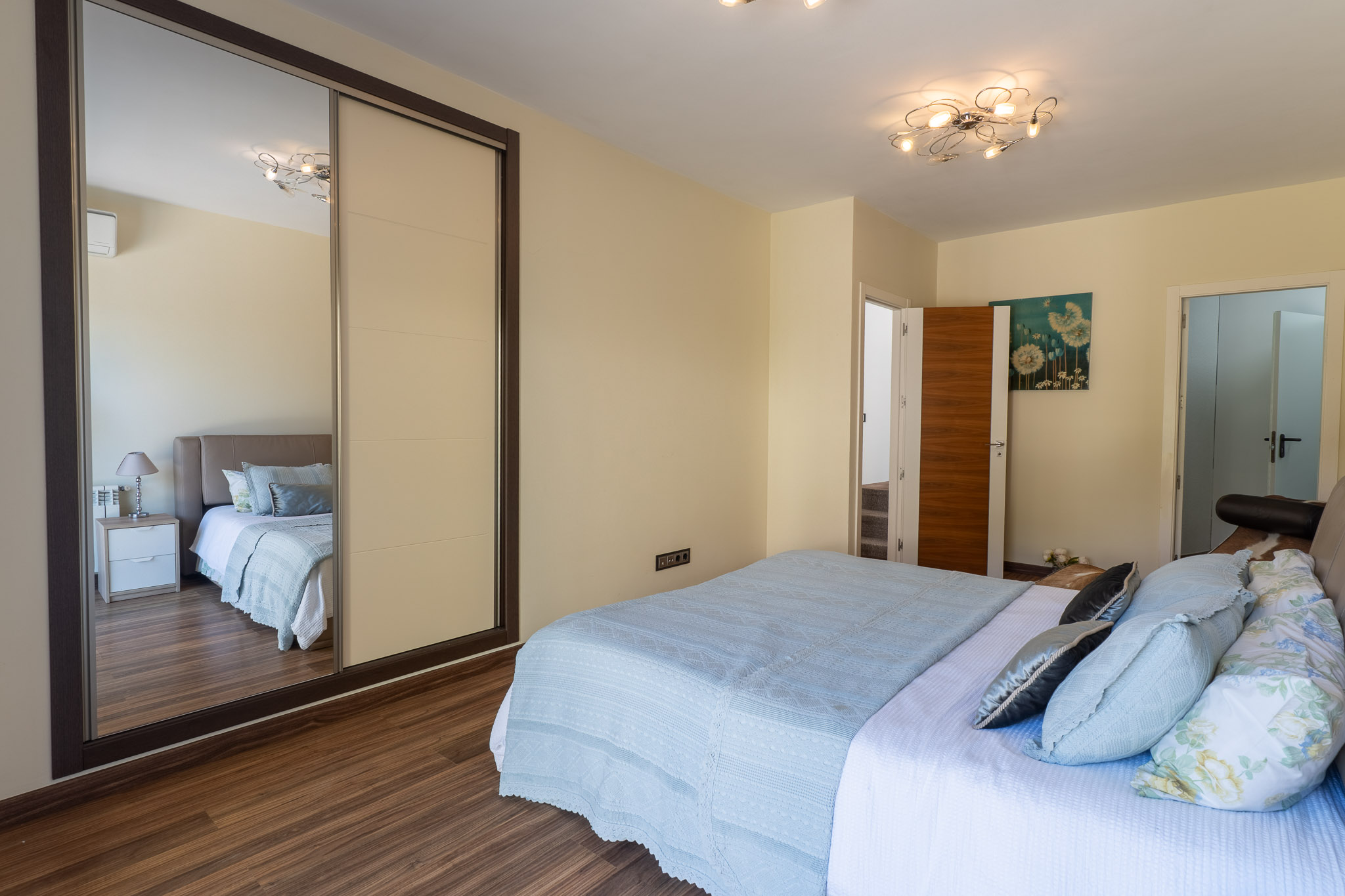 Schlafzimmer mit Bett in hellblauen Textilien, Holzlaminatboden und Schiebetürenschrank mit Spiegelfront. Eine moderne Deckenleuchte und florale Wandkunst sind sichtbar.