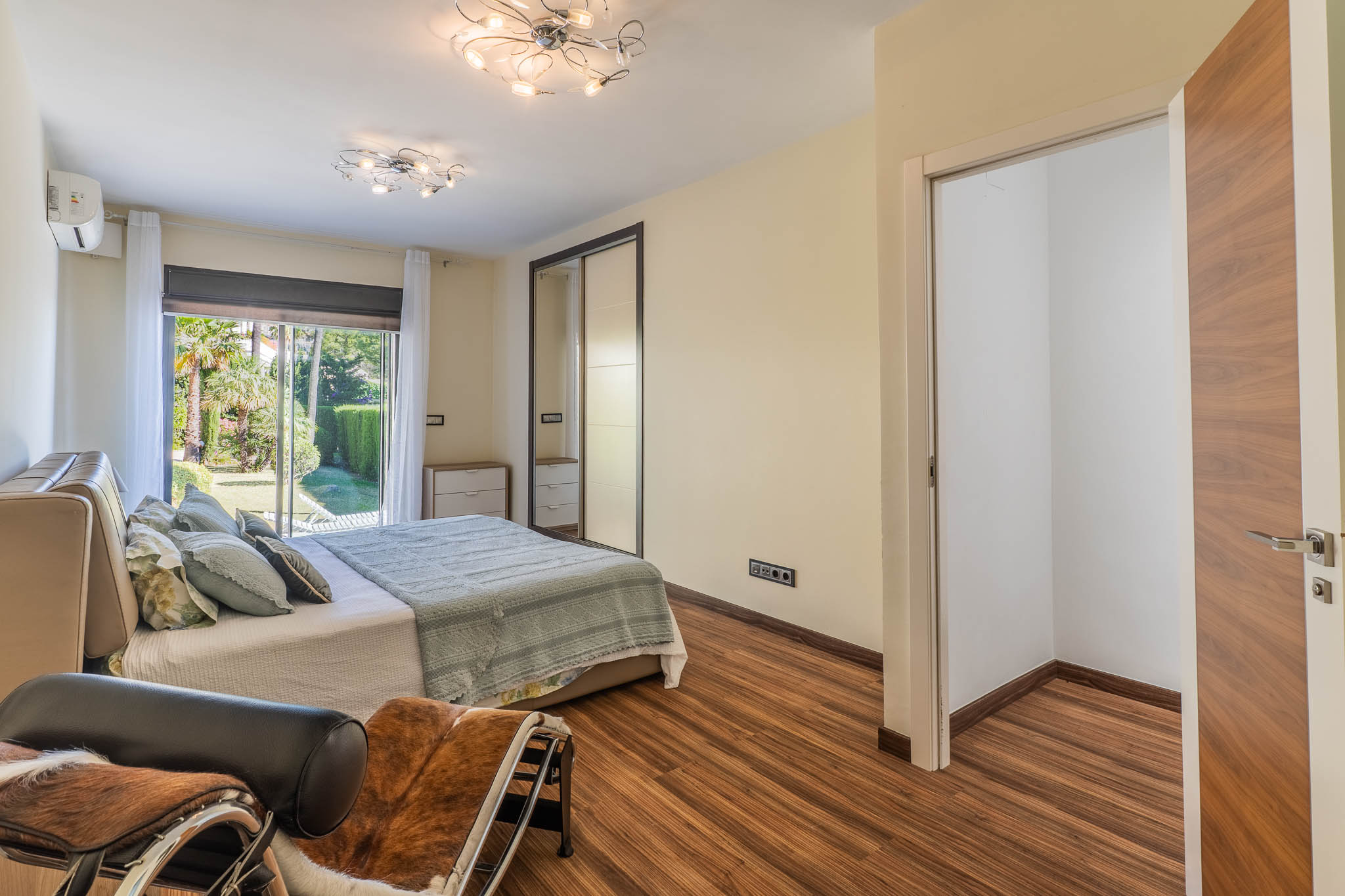 Schlafzimmer mit direktem Gartenzugang durch Glasschiebetüren, Laminatboden und Spiegelschiebetürenschrank. Ein offener Ankleidebereich ist sichtbar.