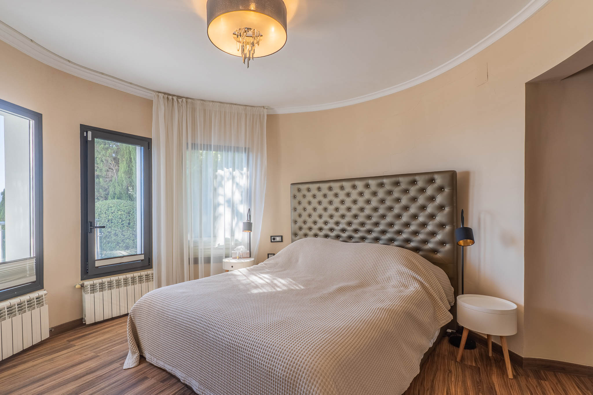 Schlafzimmer mit großem Bett und gestepptem Kopfteil. Geschwungene Wand mit dunkel gerahmten Fenstern und hellen Vorhängen. Holzboden, Heizkörper und eine moderne Deckenleuchte.