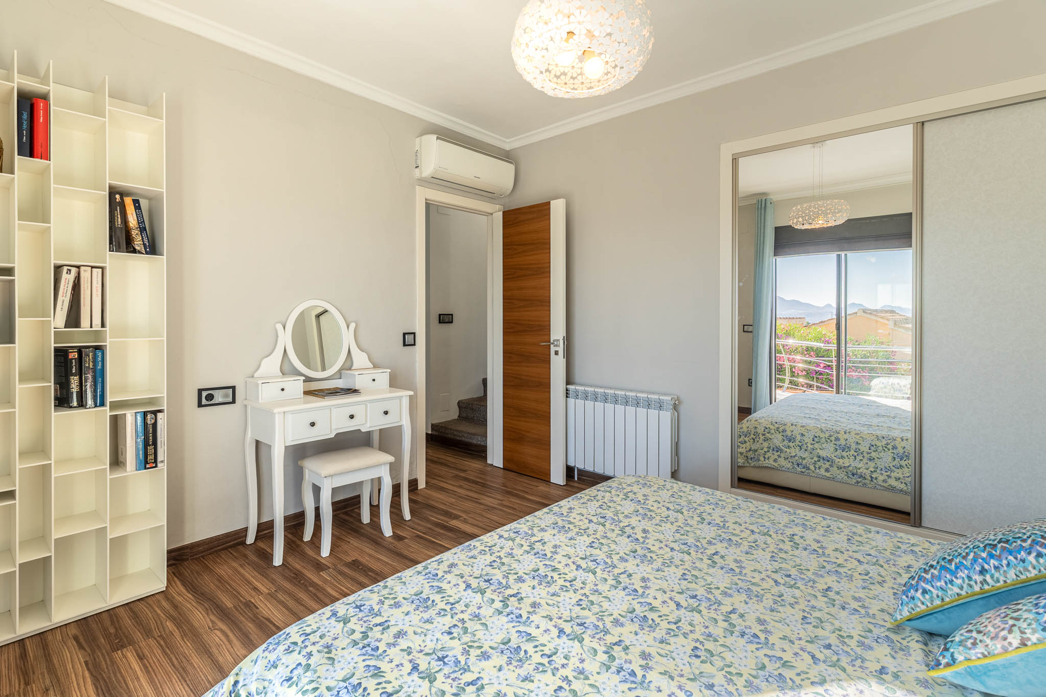 Schlafzimmer mit Laminatboden, weißem Schminktisch und modularem Bücherregal. Der Spiegelschrank reflektiert einen Bergblick vom Balkon.