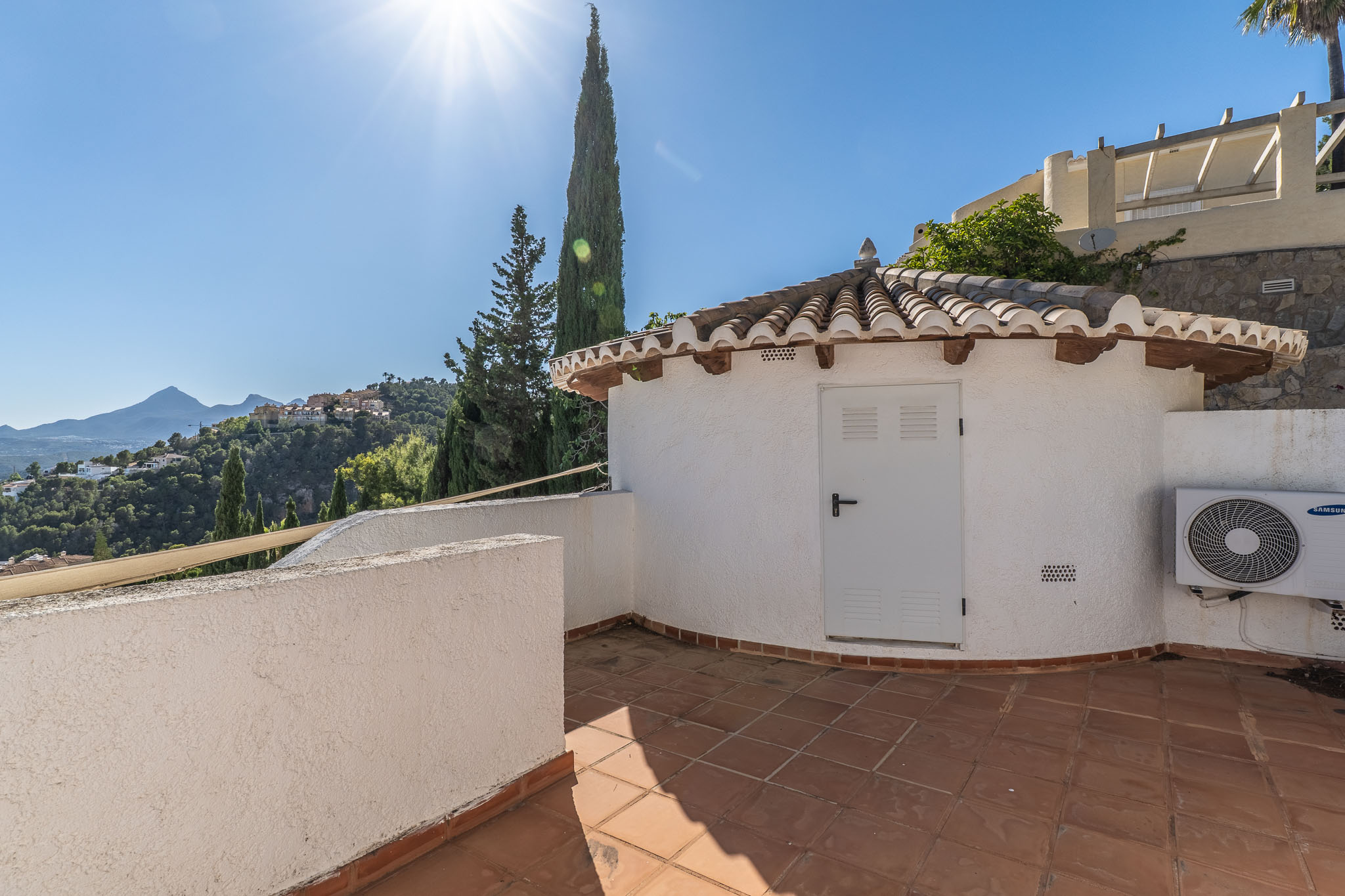 Terrasse mit Terrakottafliesen und einem charmanten Nebengebäude mit traditionellem Ziegeldach. Bietet weitreichende Ausblicke auf grüne Hügel und ferne Berge unter klarem Himmel.