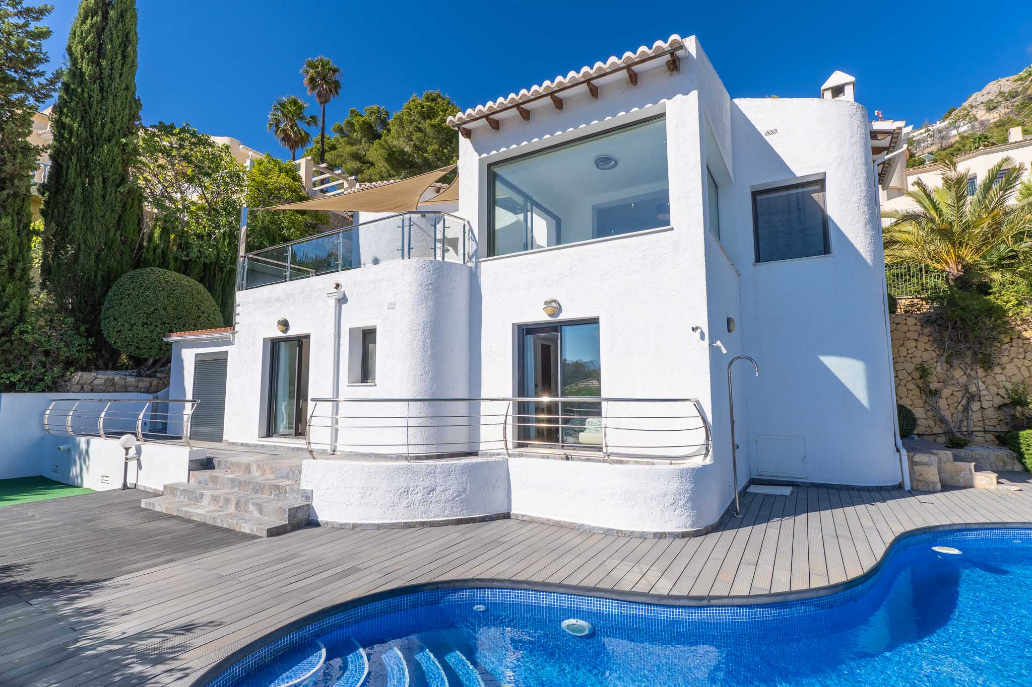 Weiße Villa mit großen Fenstern und verglasten Terrassen. Der blaue Mosaikpool und die graue Verbundterrasse werden von Zypressen und Palmen umrahmt.