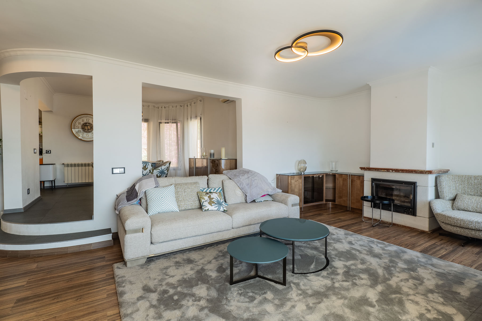 Wohnzimmer mit Kamin und rötlichem Marmorsims sowie moderner Deckenleuchte. Ein helles Sofa und tealfarbene Couchtische auf grauem Teppich führen zu einem erhöhten Bereich.