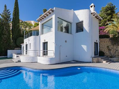 Außenansicht der Villa mit geschwungenem blauen Mosaikpool, grauer Verbundterrasse, Außendusche und mediterraner Bepflanzung.