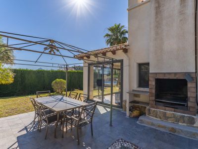 Außenterrasse mit dunkelgrauen Fliesen, Metallpergola und Essgruppe. Ein gemauerter Grill aus Ziegeln und Stein sowie Glasschiebetüren zum Innenbereich sind sichtbar.